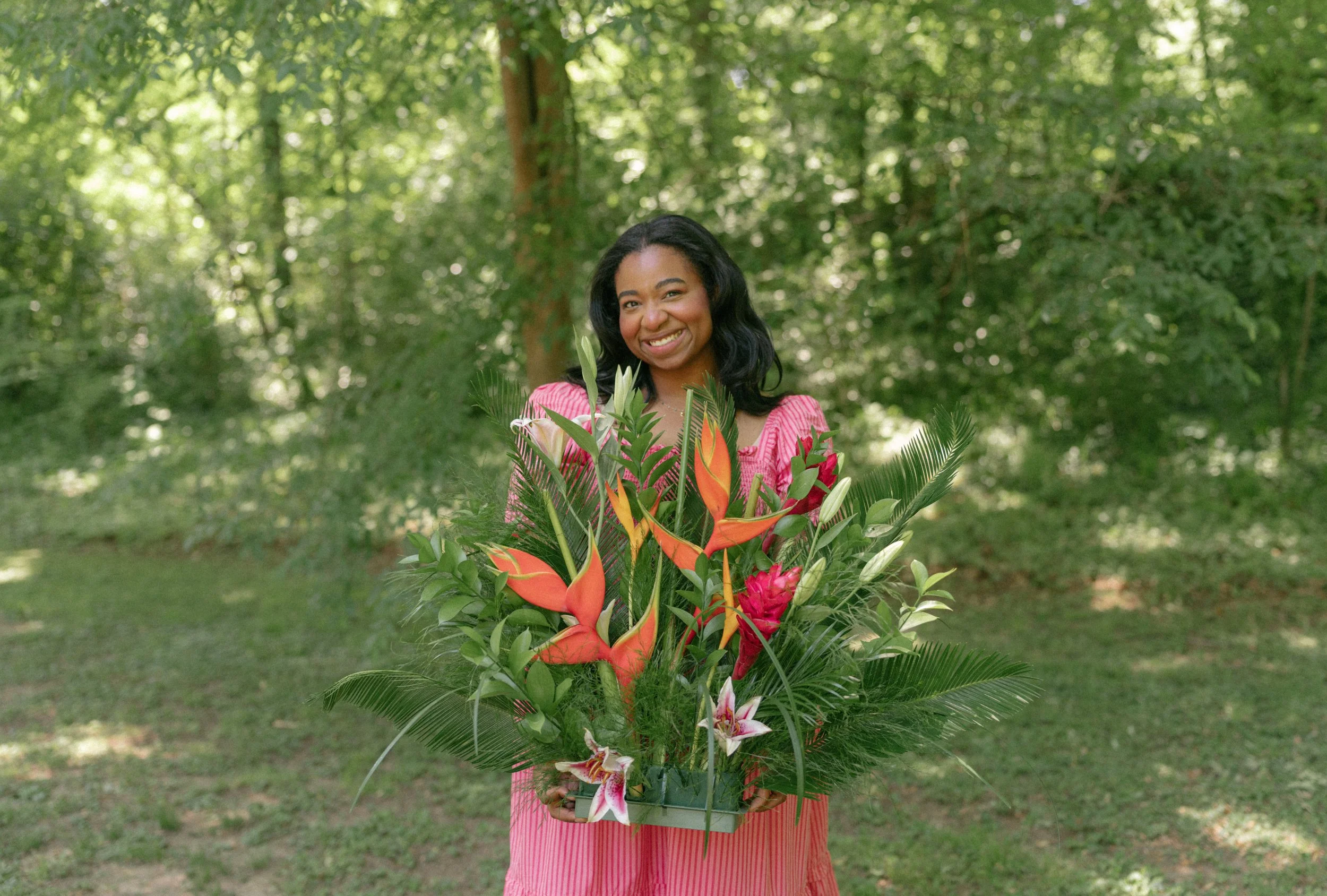 A woman wearing a pink dress holding a large bouquet of colorful flowers outdoors surrounded by green trees