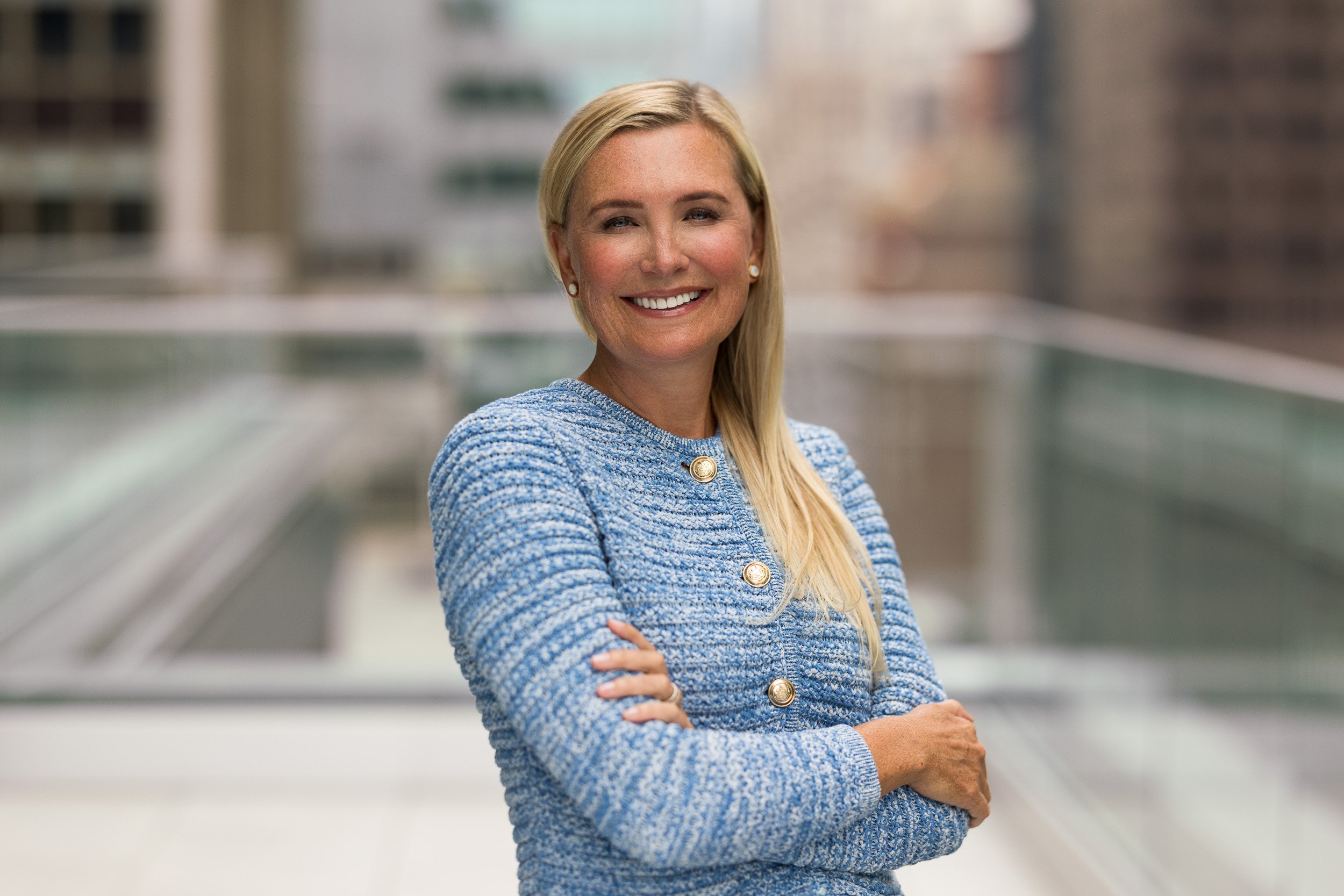 Modern corporate headshot of a female CEO wearing a blue sweater and smiling outdoors on the terrace of her office with a view of the NYC skyline