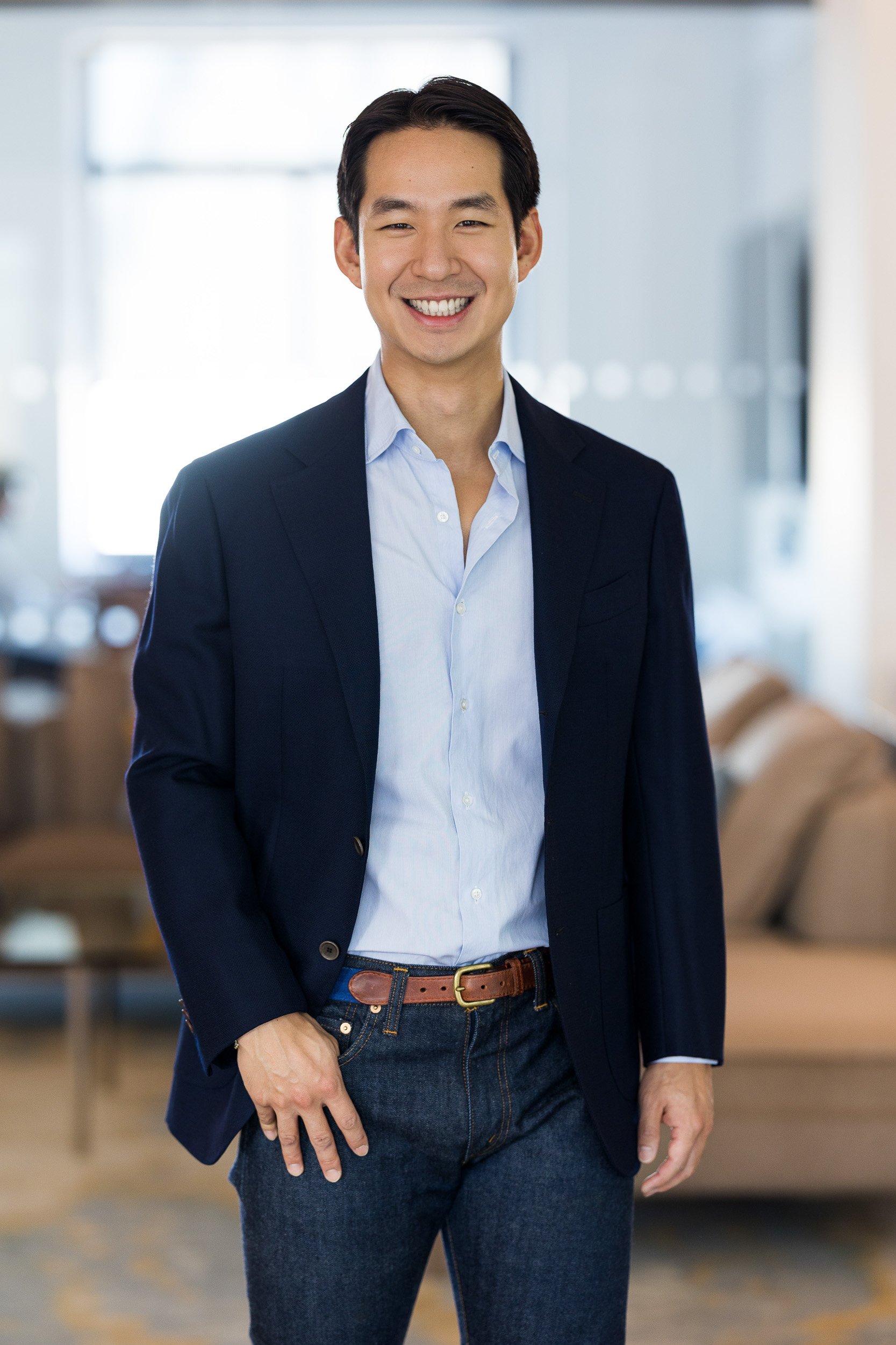 Corporate headshot of Asian male wearing a navy blazer and light blue dress shirt and dark blue jeans with a brown belt featured against a blurred office background.