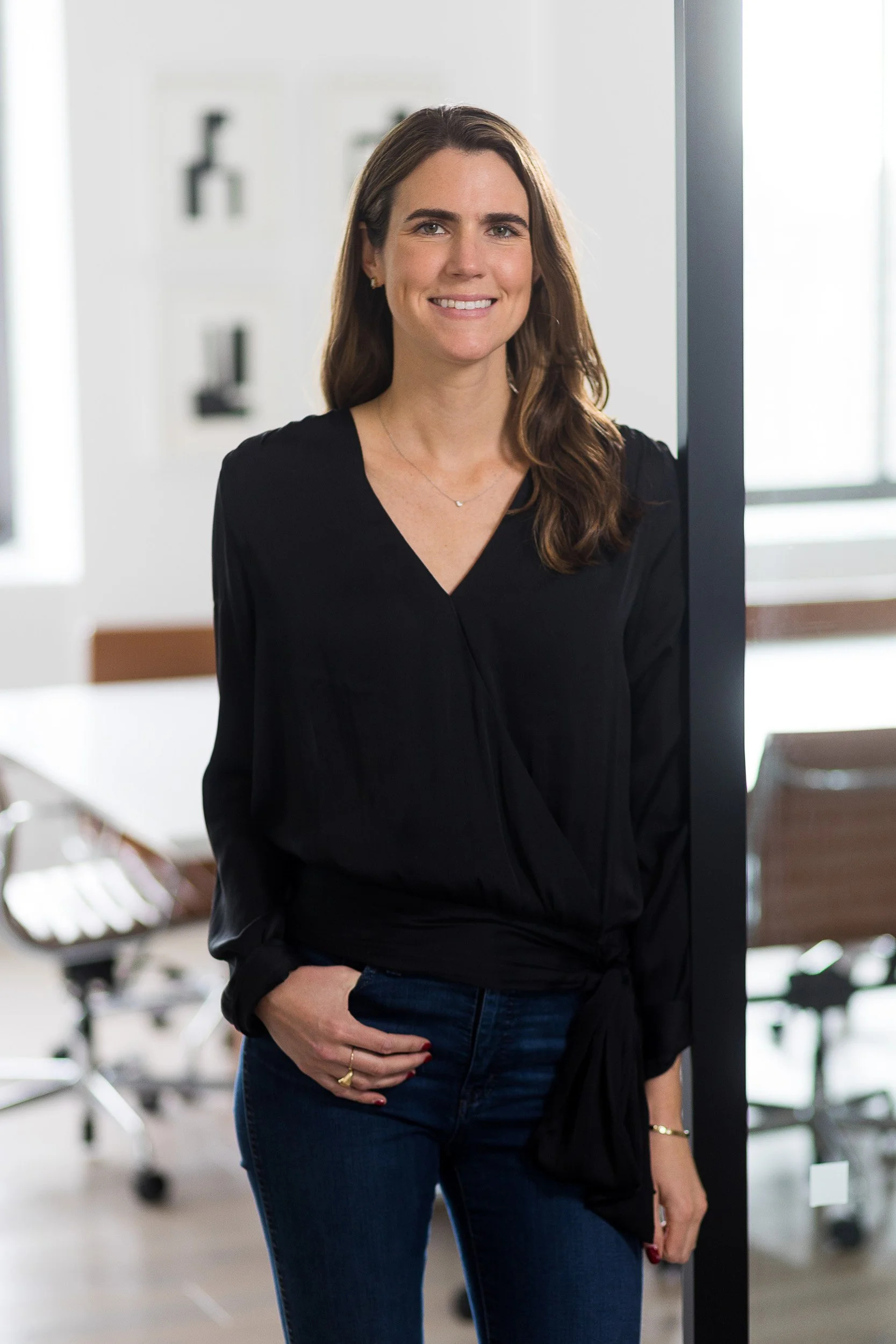 Corporate portrait of a female in her 30s wearing a black blouse and blue jeans while posing in an office