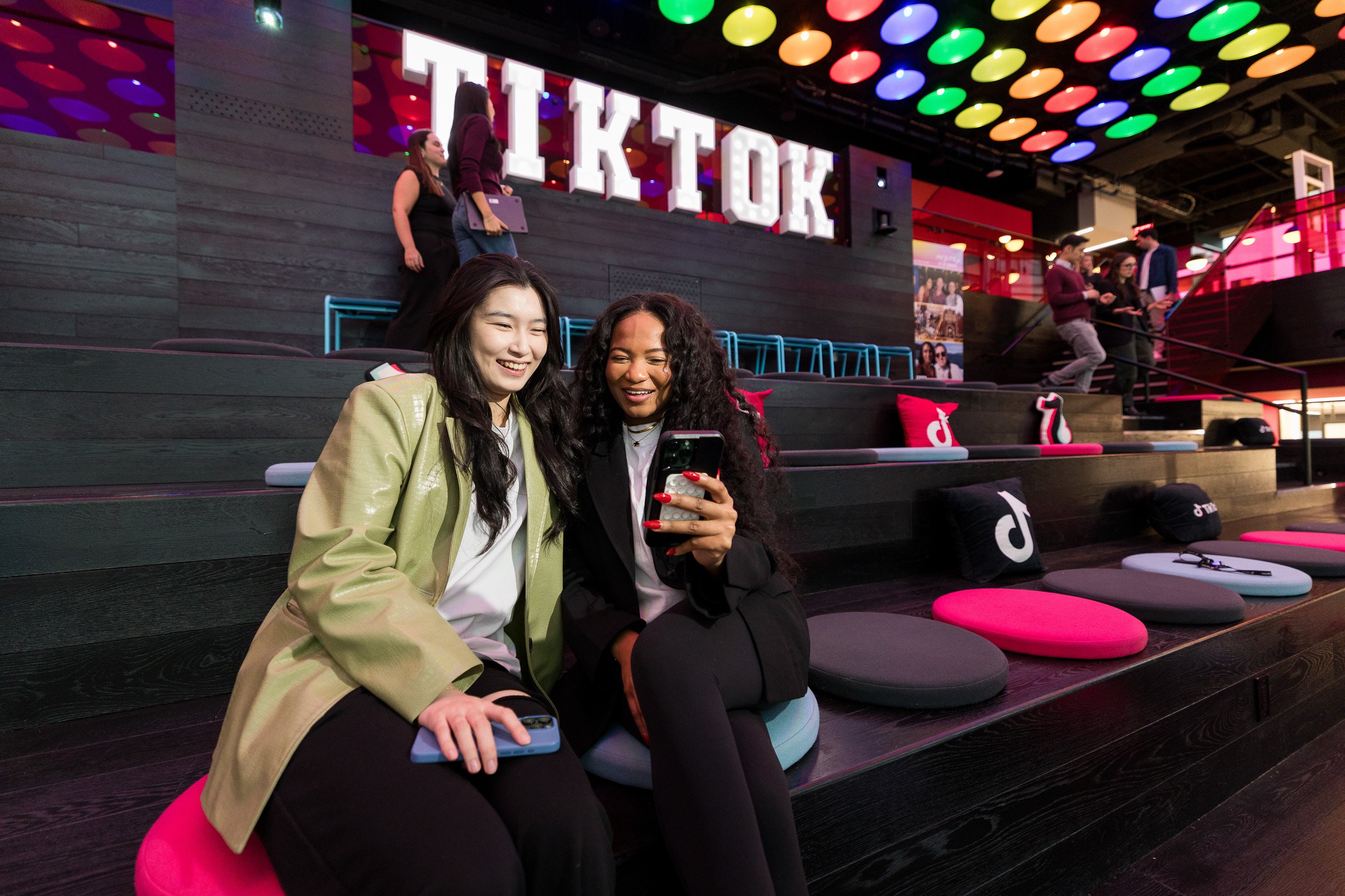 Two young women sitting in a large colorful room looking at a phone with a large sign that says TikTok in the background.