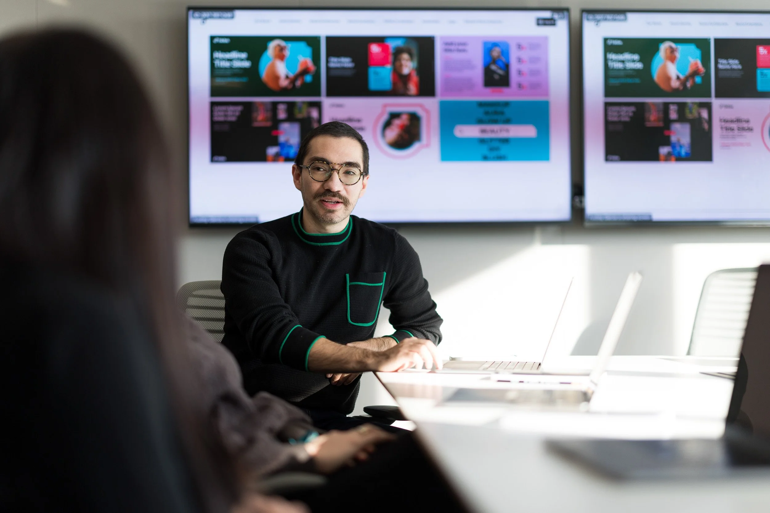 Corporate lifestyle photography of a man sitting at a conference room table with laptops on the table and tv screens in the background featuring company marketing materials. 