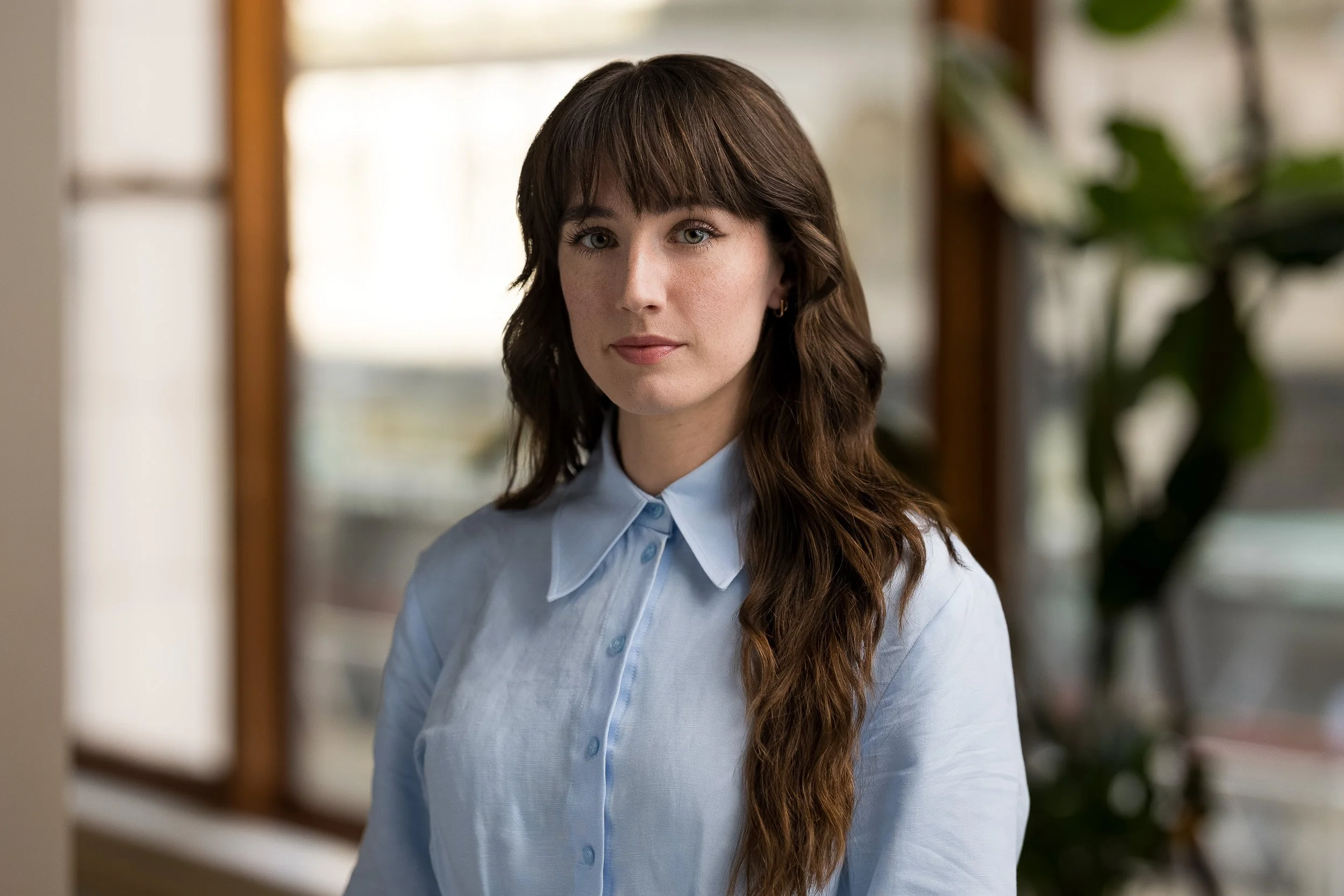 Modern corporate headshot of young white female wearing a blue blouse with a serious expression in an office environment