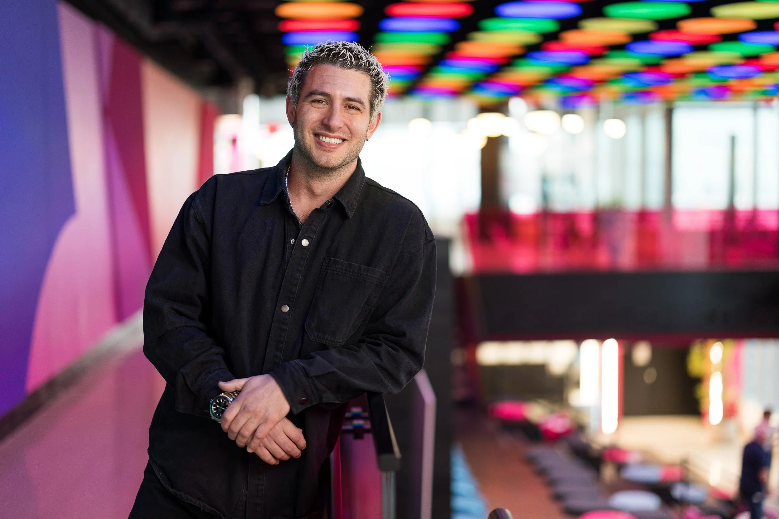 Creative corporate headshot of young white male leaning against a handrail in a large colorful office