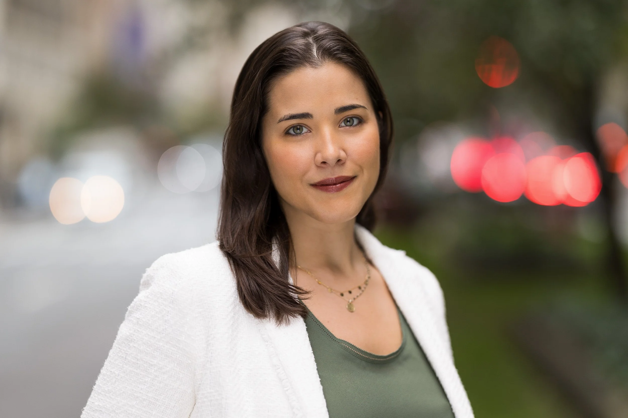 Professional headshot of female in her 30's wearing a white sweater-blazer and green blouse outdoors in NYC