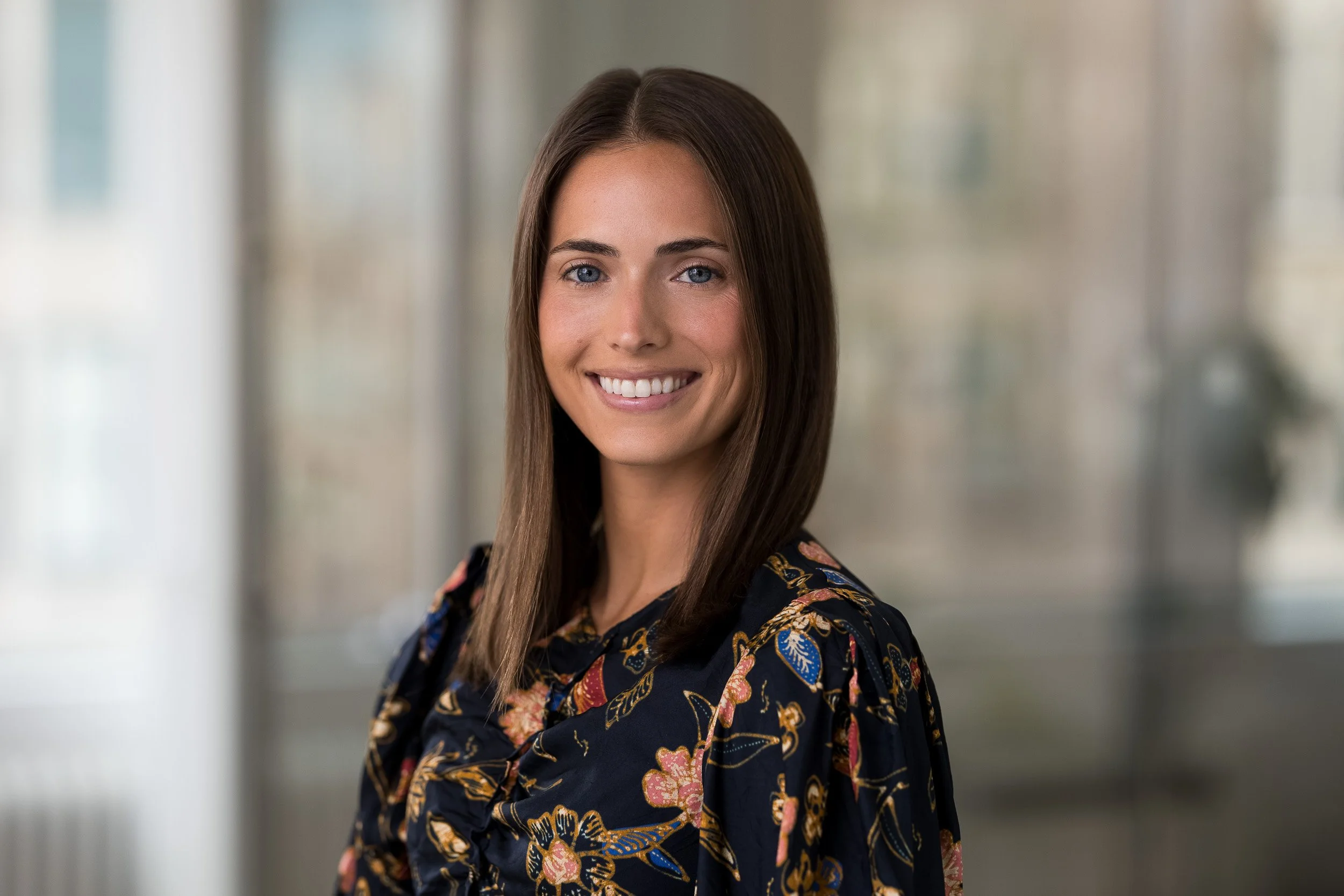 Female orporate headshot of a smiling young white woman with long brown hair in a floral top posing in her NYC office