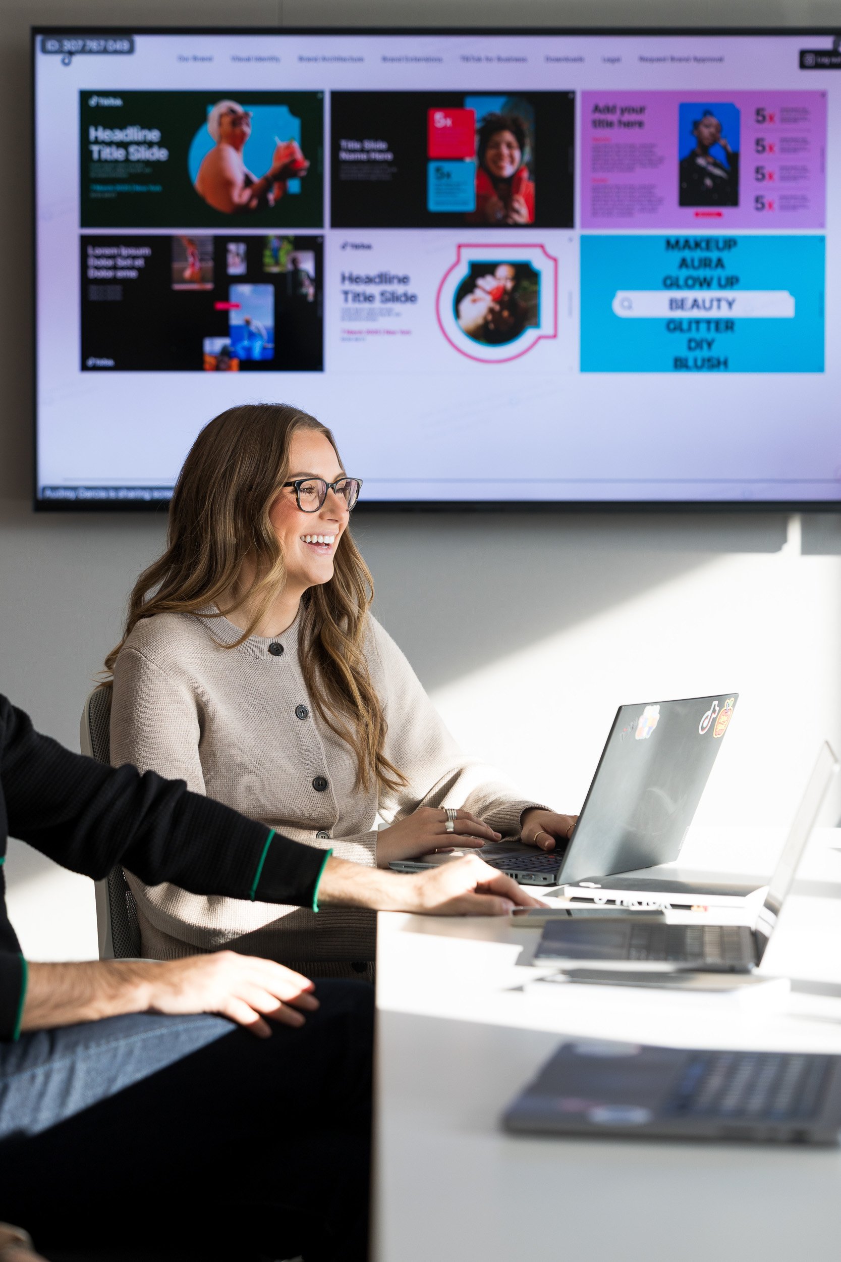 Corporate lifestyle photography of a young woman sitting at a conference room table smiling in front of her laptop with a large screen in the background featuring company marketing materials.