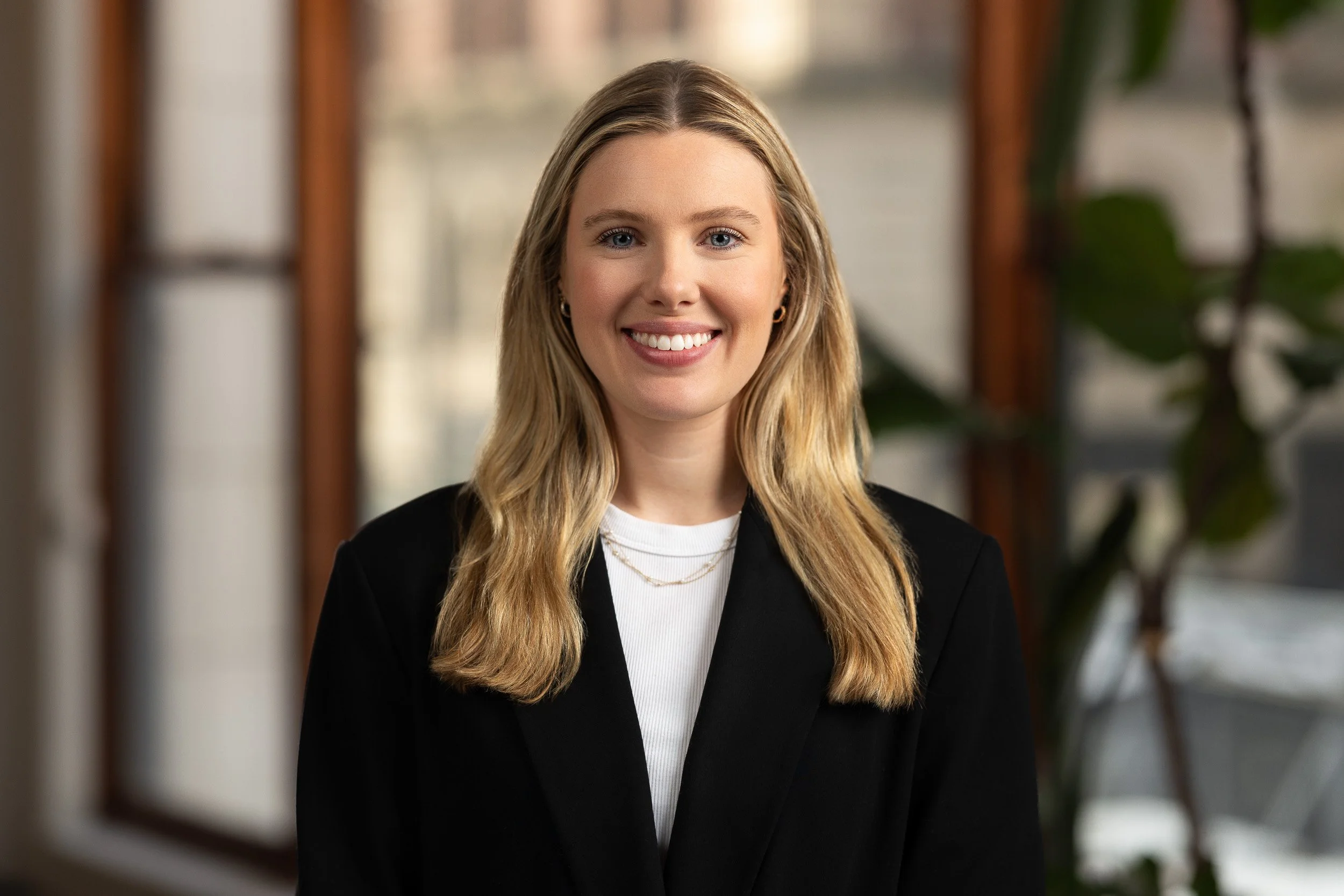 Environmental headshot of a blonde woman wearing a black blazer and white blouse photographed indoors with a sunny window scene in the background