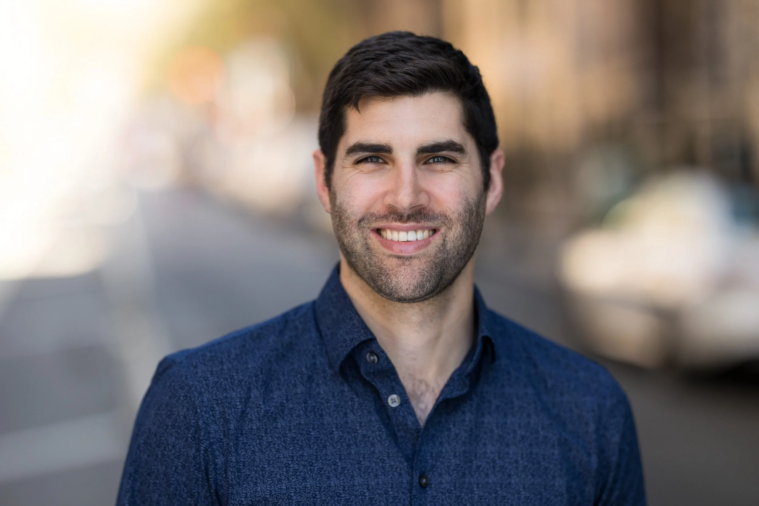 Professional headshot of young white male employee wearing a textured dark blue dress shirt outdoors in NYC.
