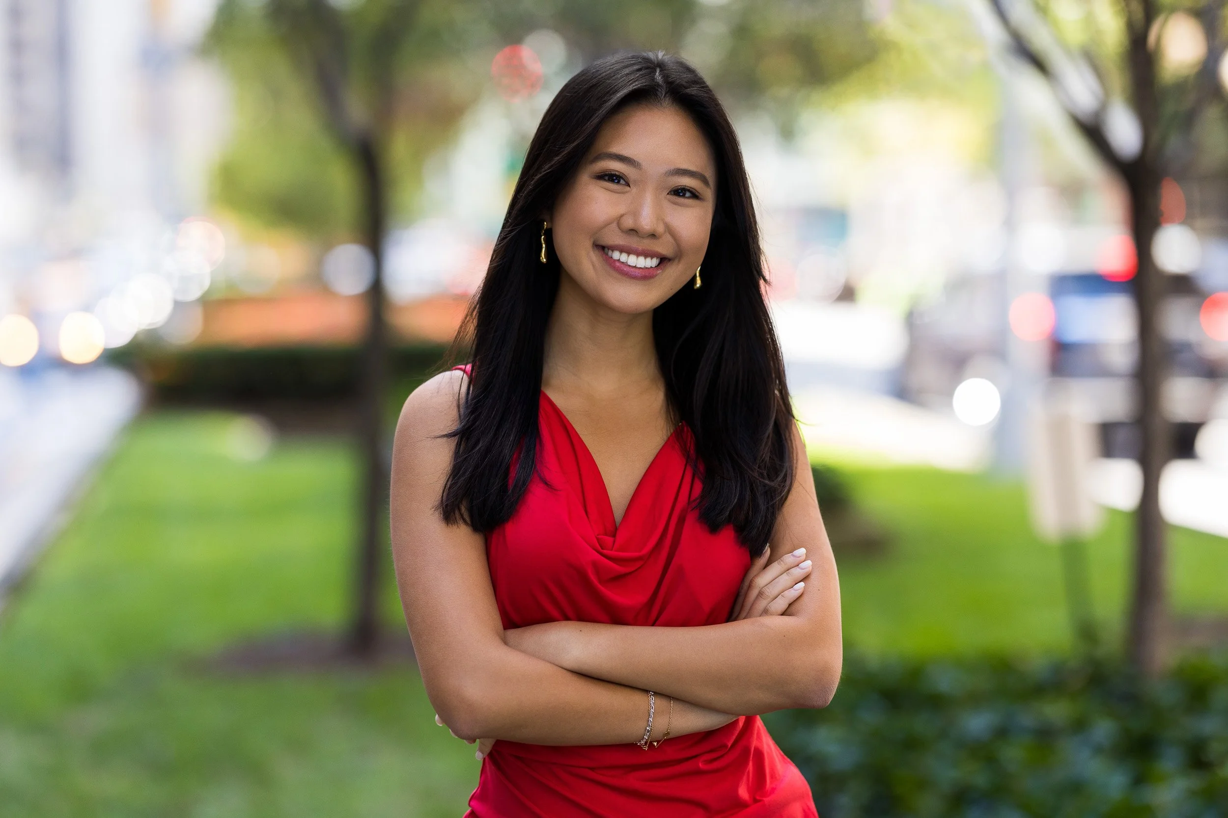 Modern corporate headshot of a smiling young Asian woman with long black hair in a red sleeveless dress standing outdoors in NYC with arms crossed, blurred city street and trees in the background.