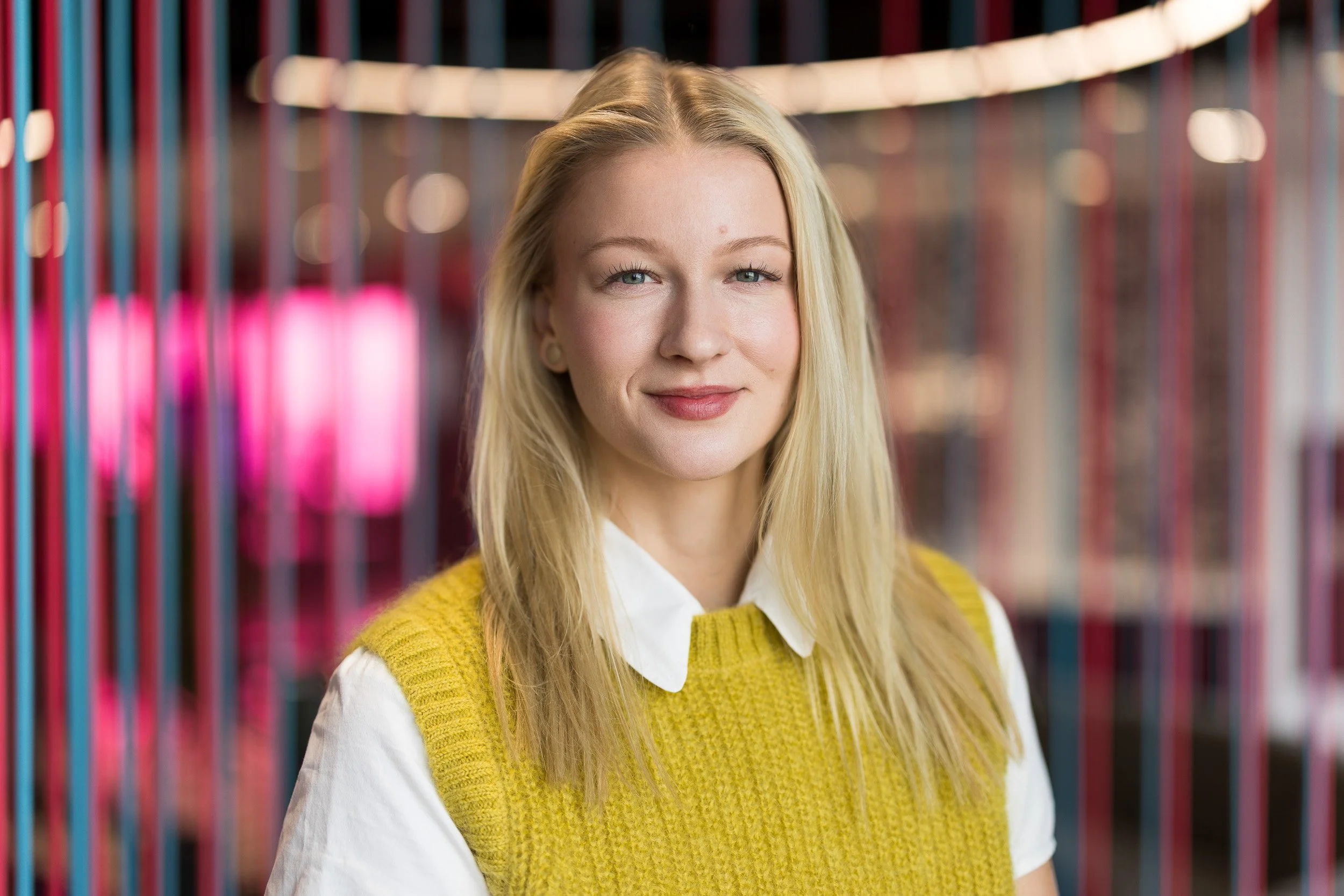 Creative corporate headshot of young white female employee wearing a yellow sweater vest and white blouse with light blonde hair smiling in a colorful NYC office setting.