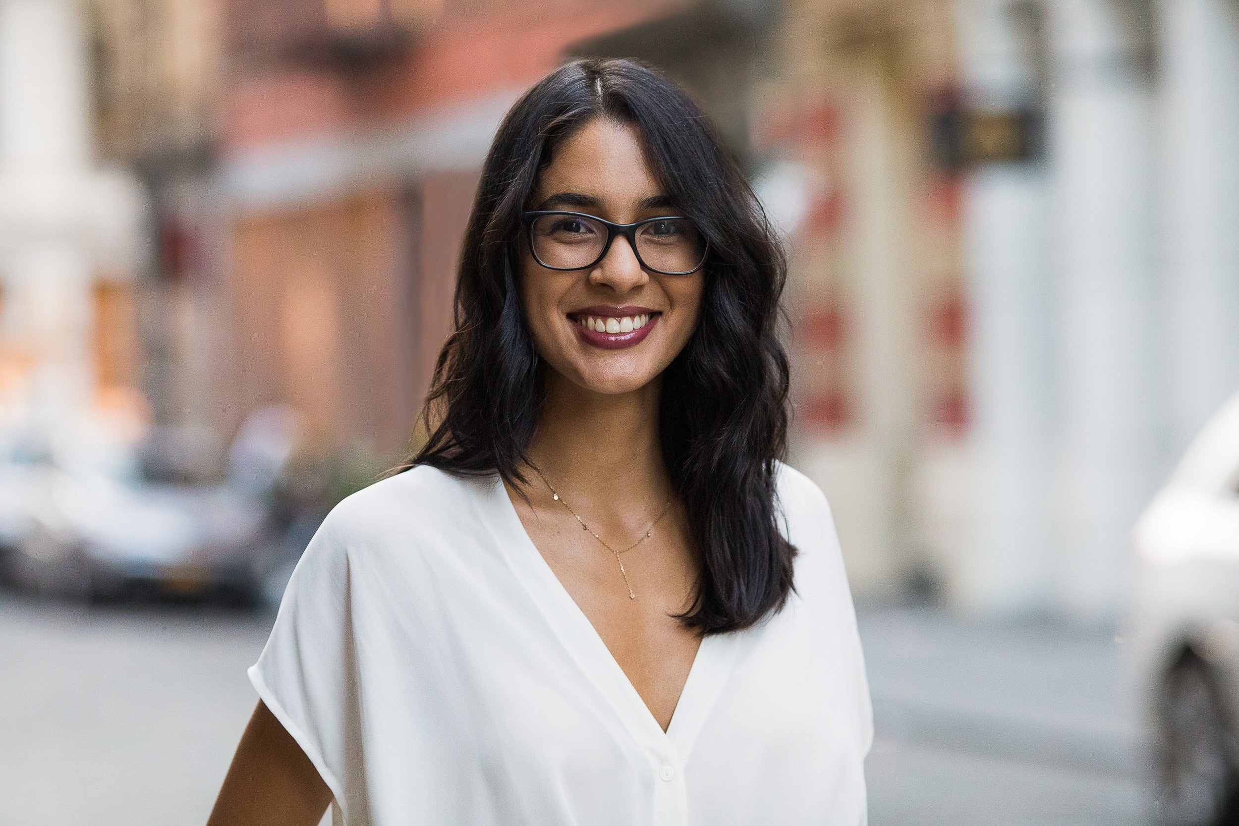 Female professional headshot of young Indian woman wearing a white blouse and glasses smiling outdoors in NYC