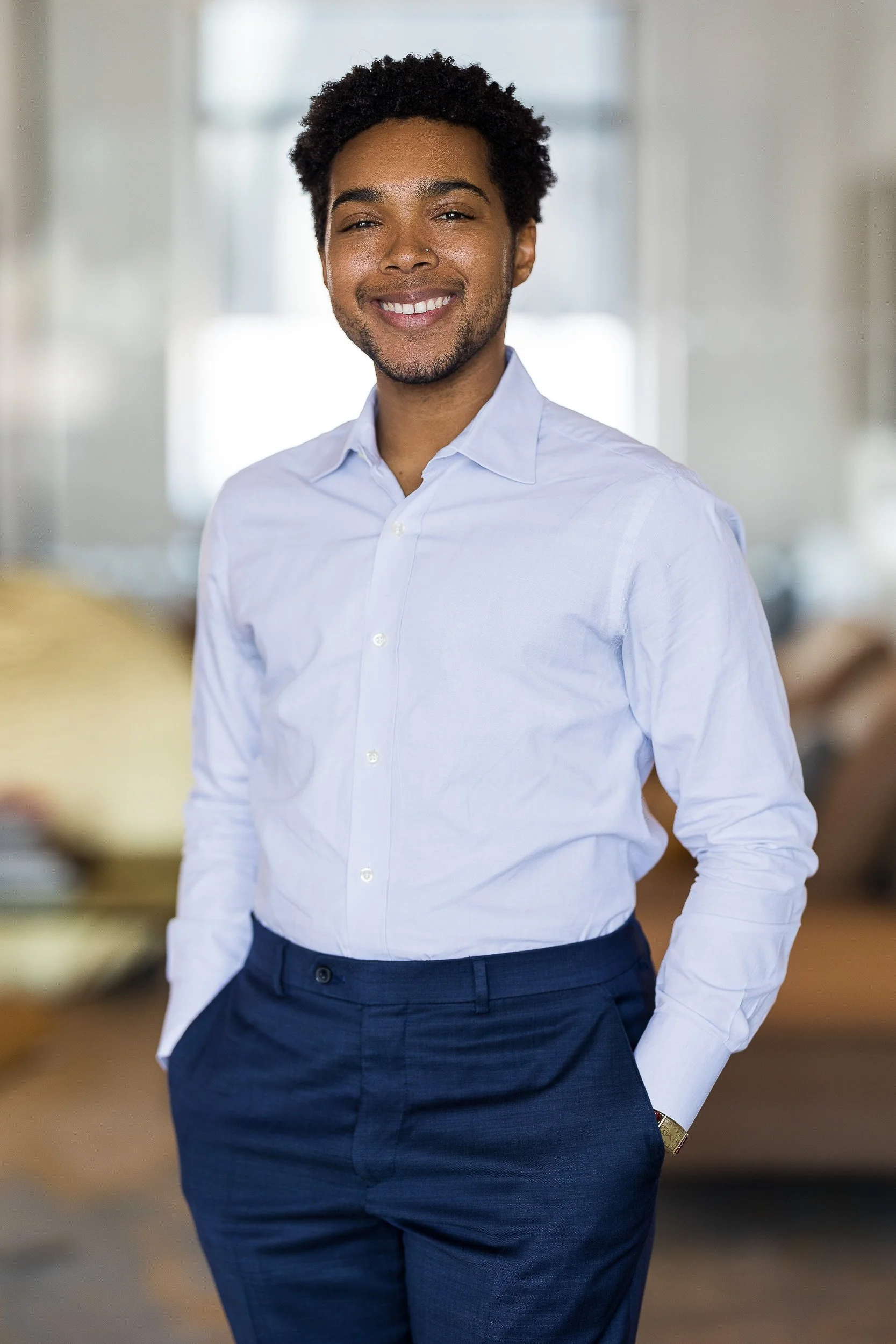Modern professional portrait of male in his 20's wearing a blue dress shirt and navy dress pants with a blurred office background