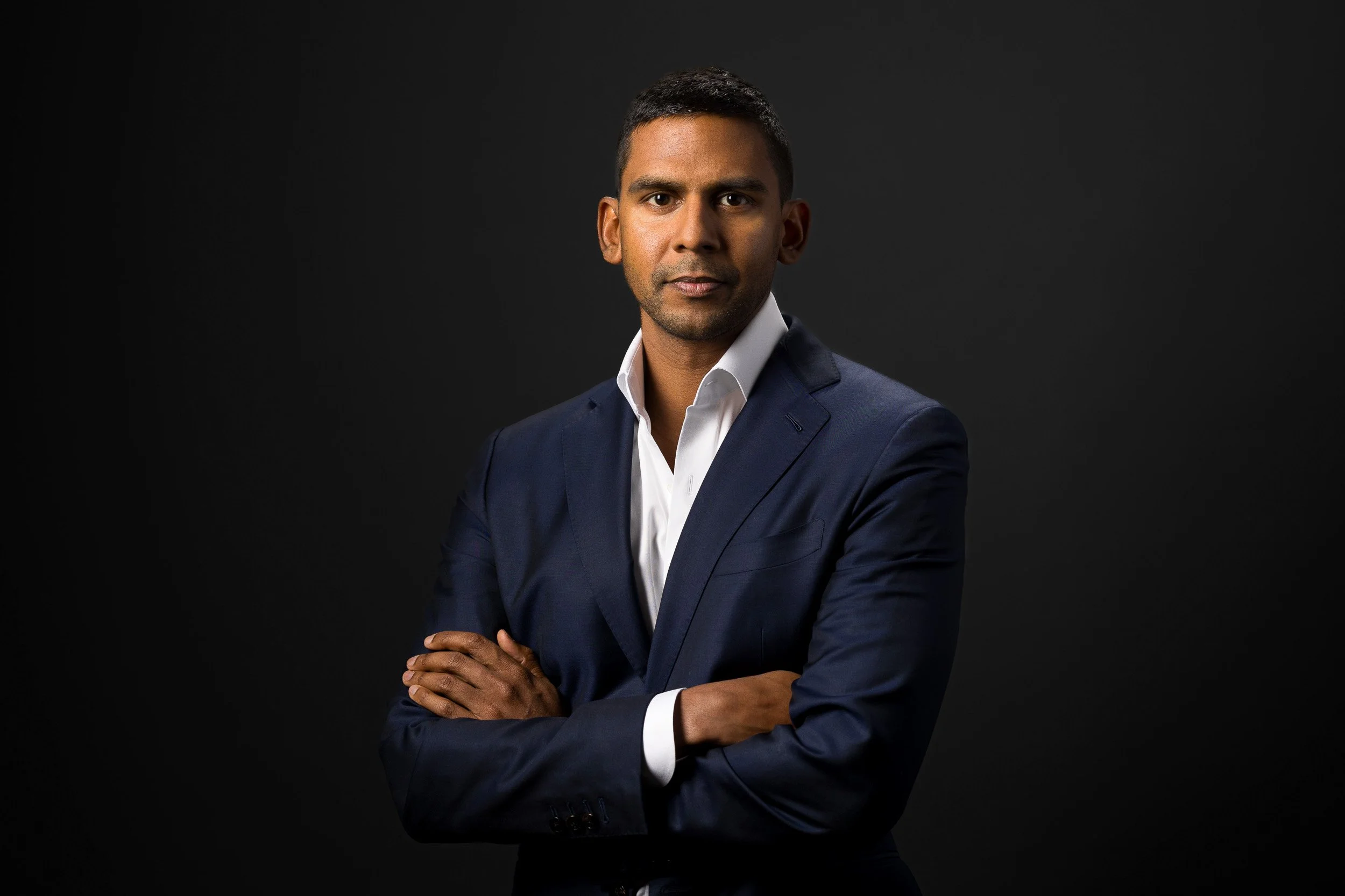 Corporate headshot of middle-aged Indian male in navy suit and white shirt against a black studio background