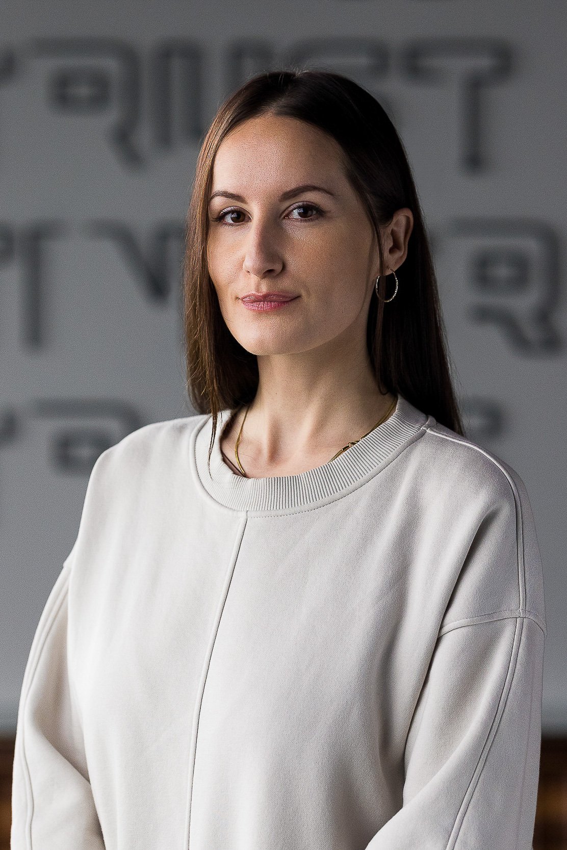 Creative corporate headshot of young white female employee with brown hair and earrings wearing an oversized gray sweatshirt against a blurred gray wall with writing on it.