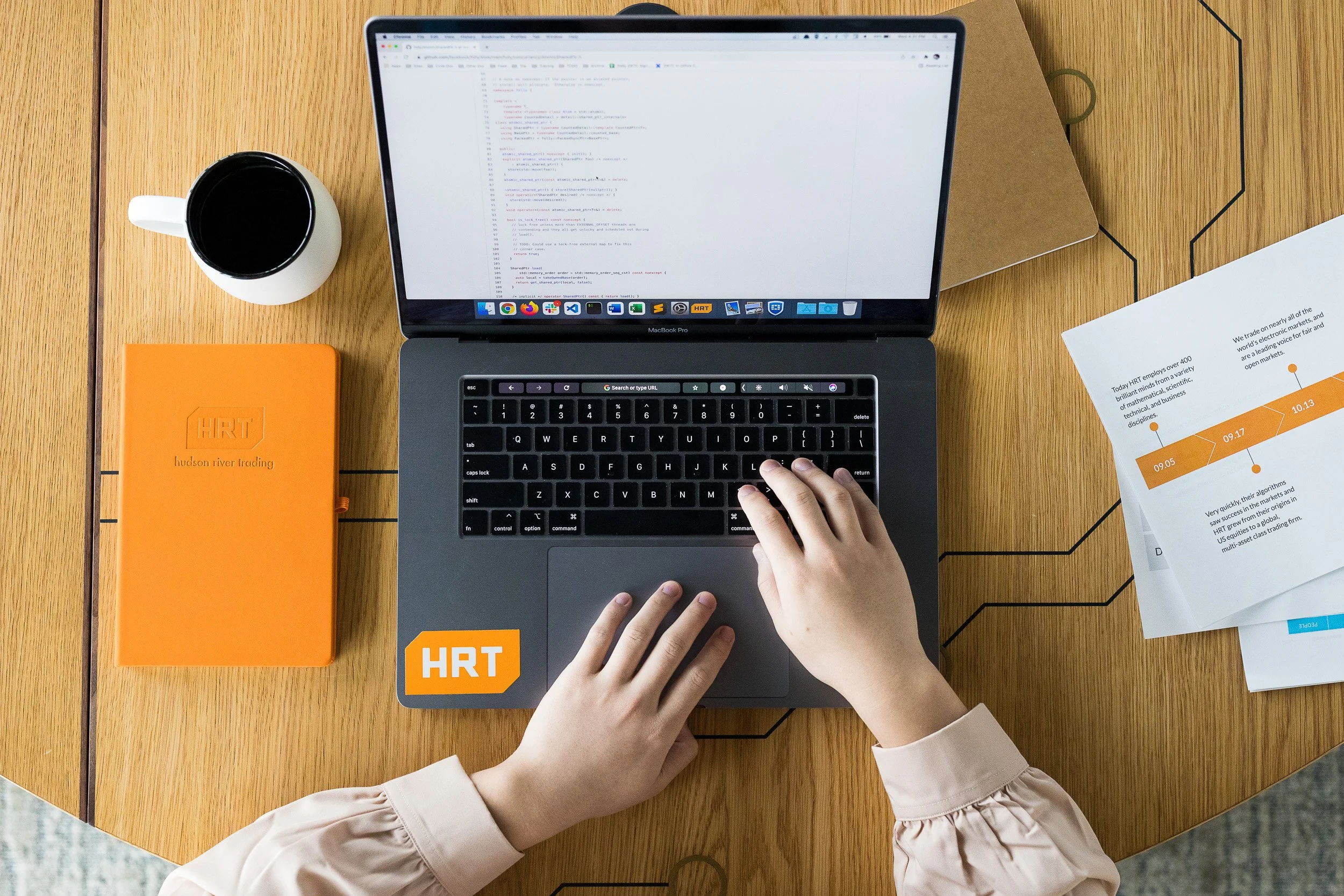 Corporate lifestyle photography overhead image of a person's hands using a MacBook Pro laptop on a wooden desk, with a coffee mug, an orange company-branded notebook, a brown folder, and printed documents nearby.