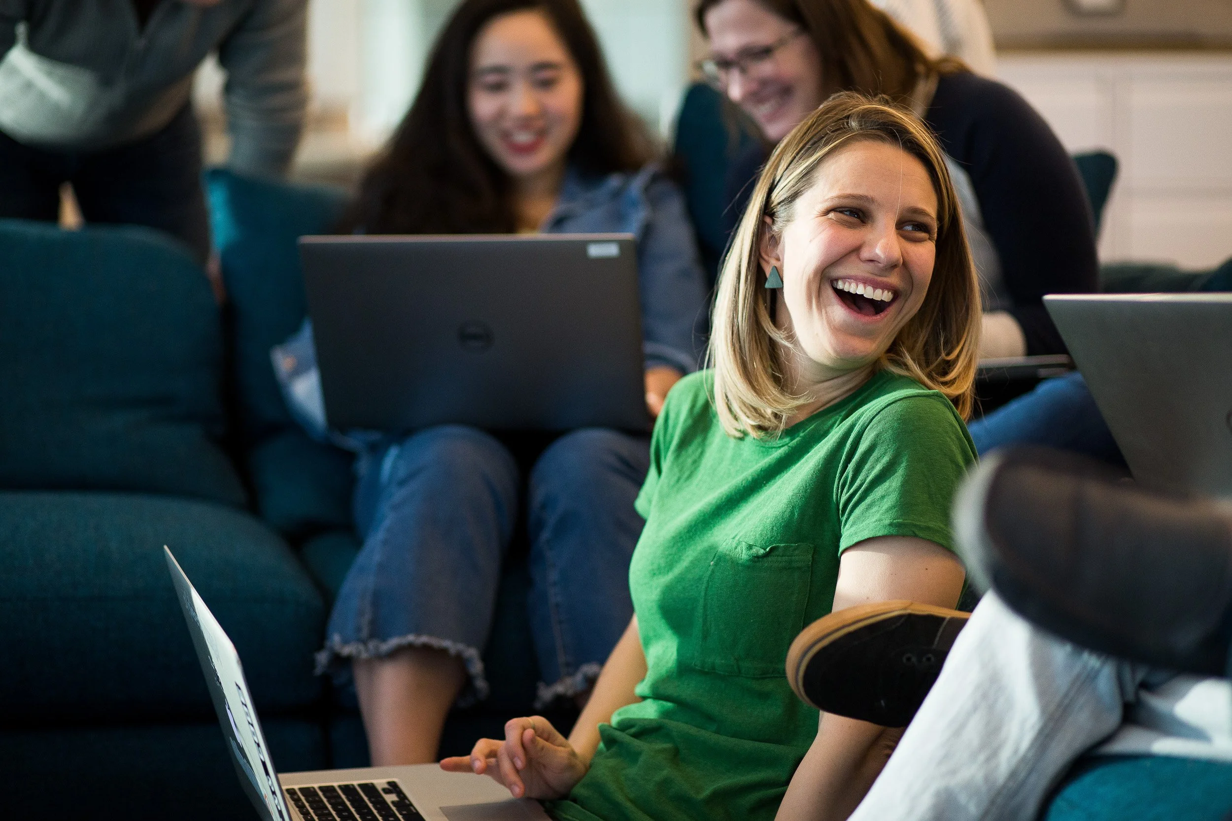 Corporate lifestyle photography of a happy woman at work in NYC. She is sitting with her laptop, with two other women laughing in the background who are both sitting on a sofa with a laptop in an office.