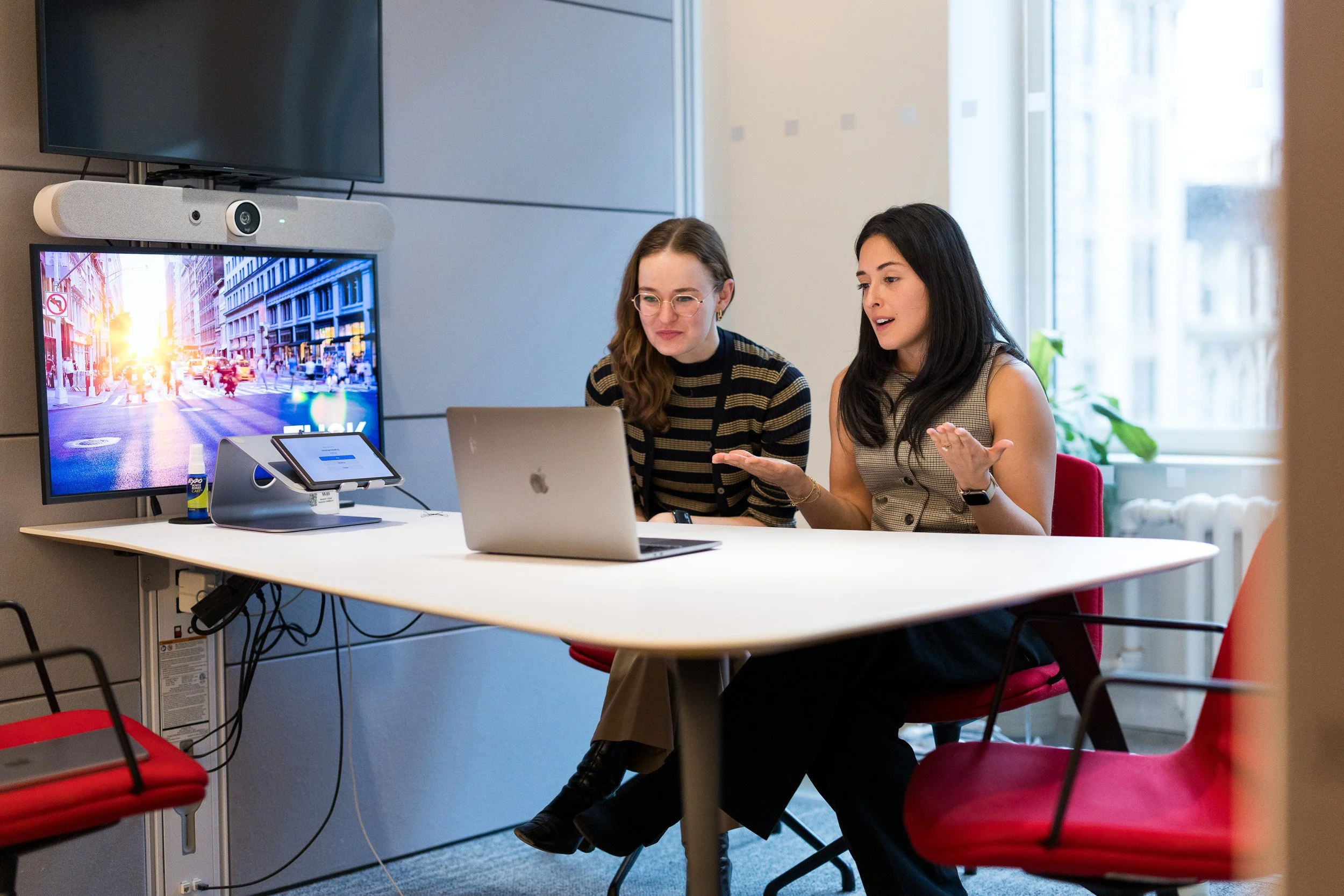 Corporate lifestyle photography of two young female employees holding a zoom call in a small meeting room in their NYC office. One is speaking and gesturing with her hands while the other listens. 