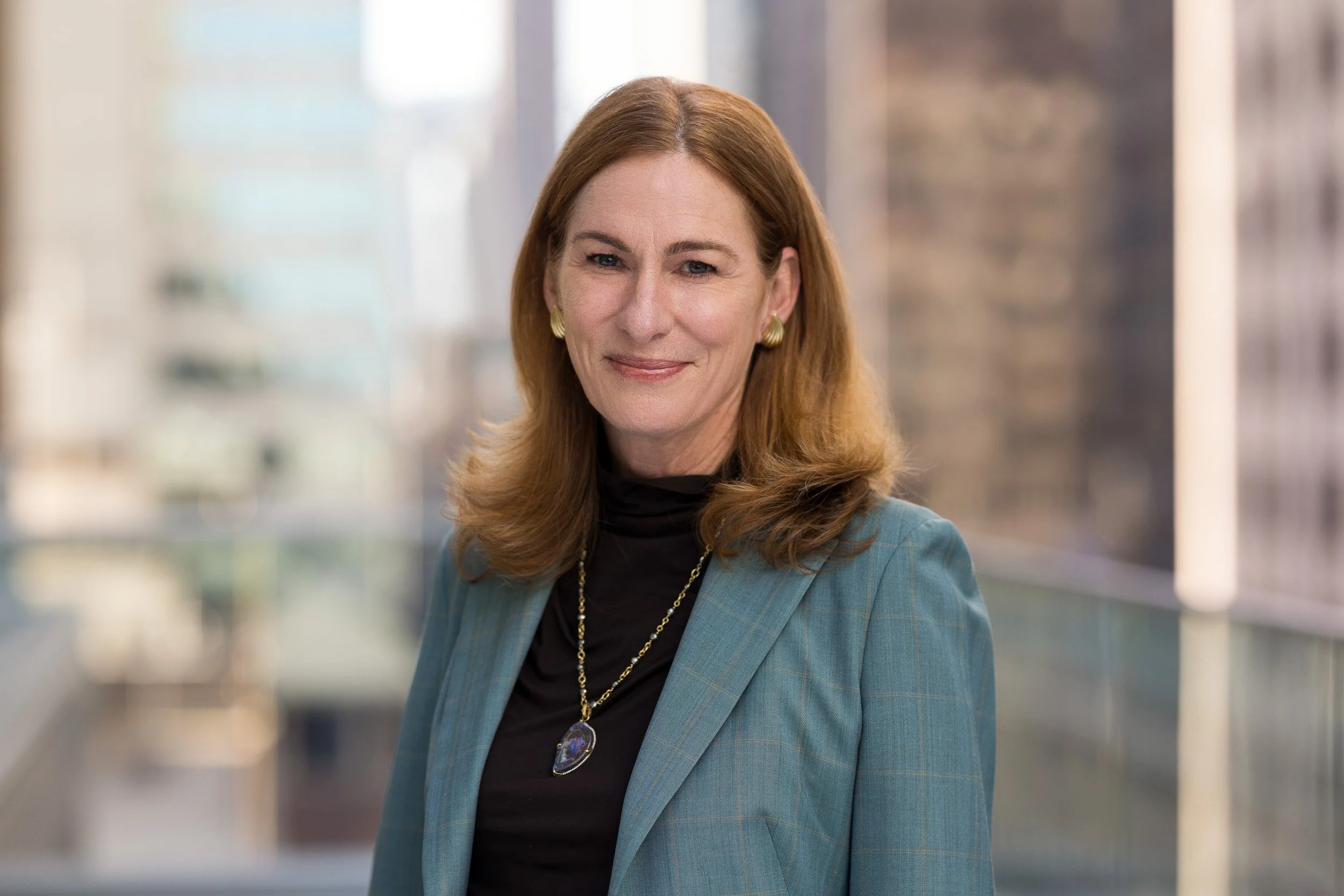 Female professional headshot of a middle-aged white woman with light brown hair wearing a blue blazer outdoors against a blurred NYC skyline background