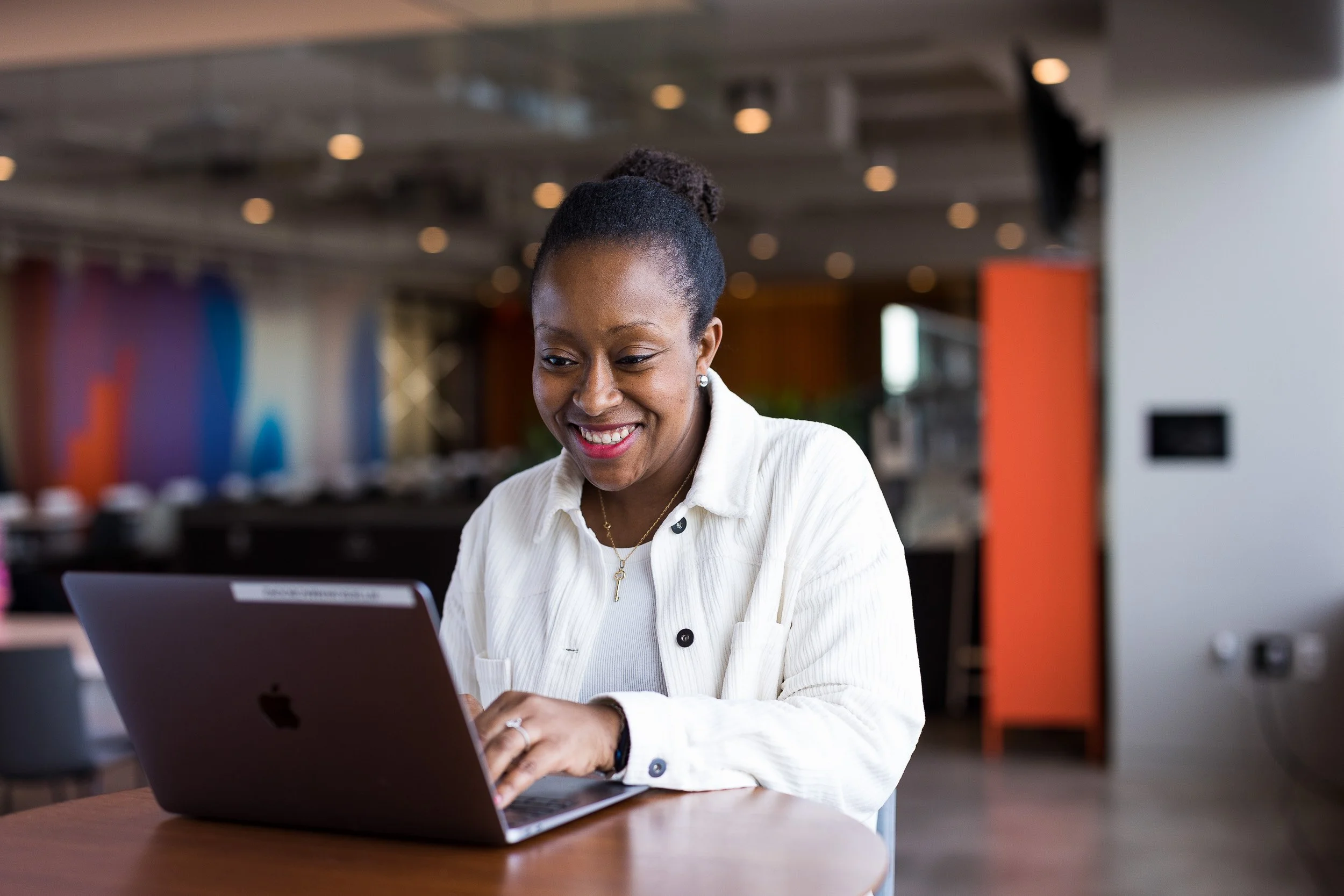 Corporate lifestyle photography of a young woman sitting at a table, smiling while working on a silver laptop in a modern, colorful cafe area of her workplace. 