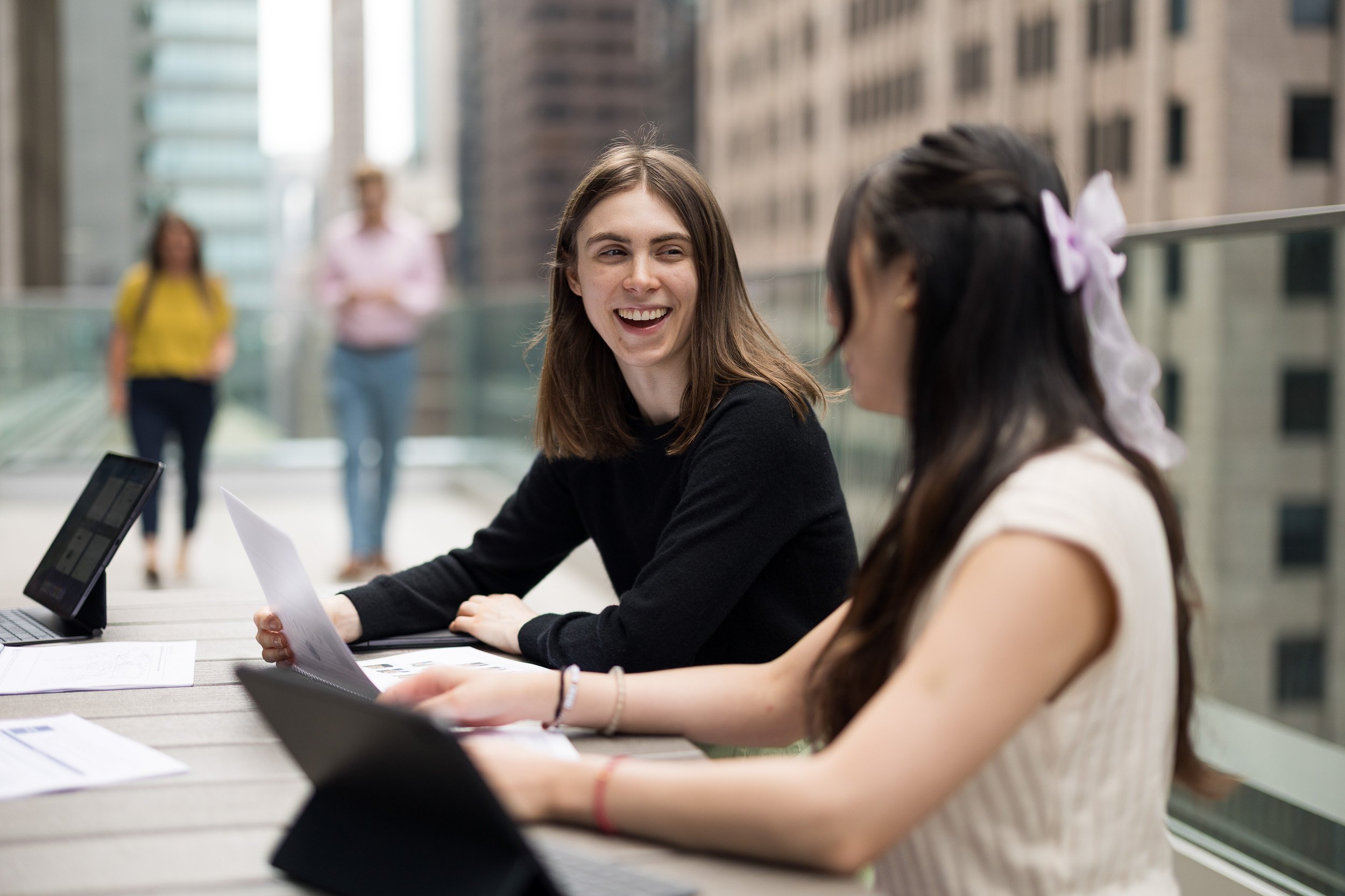 Corporate lifestyle photography of two women coworking outdoors on their office's terrace, with two blurred figures walking in the background and a view of the NYC skyline. 