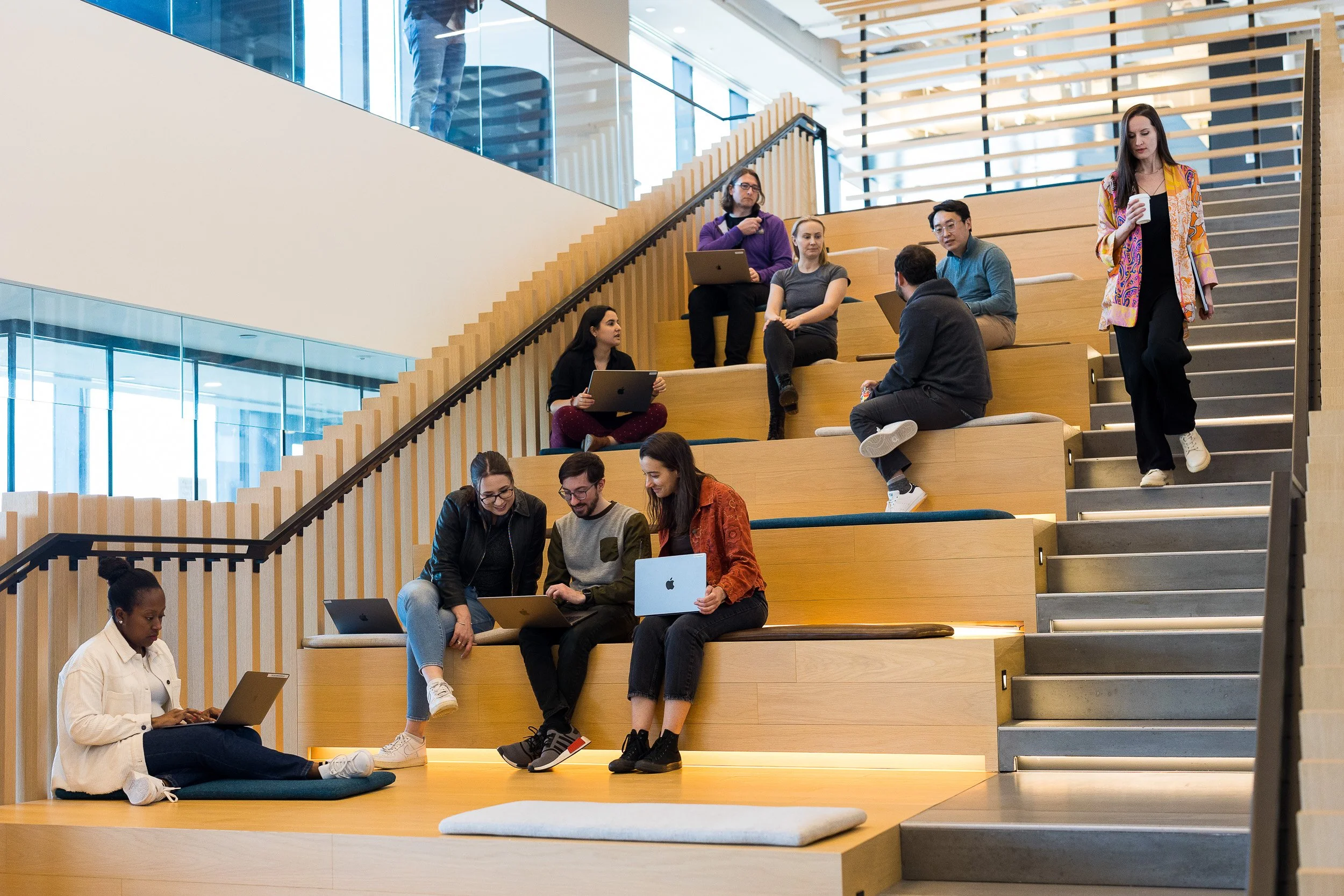 Corporate lifestyle photography of a large group of employees scattered throughout an informal seating area in a company's office. Some people are working on laptops, others are just chatting, while another person is seen walking down the stairs. 