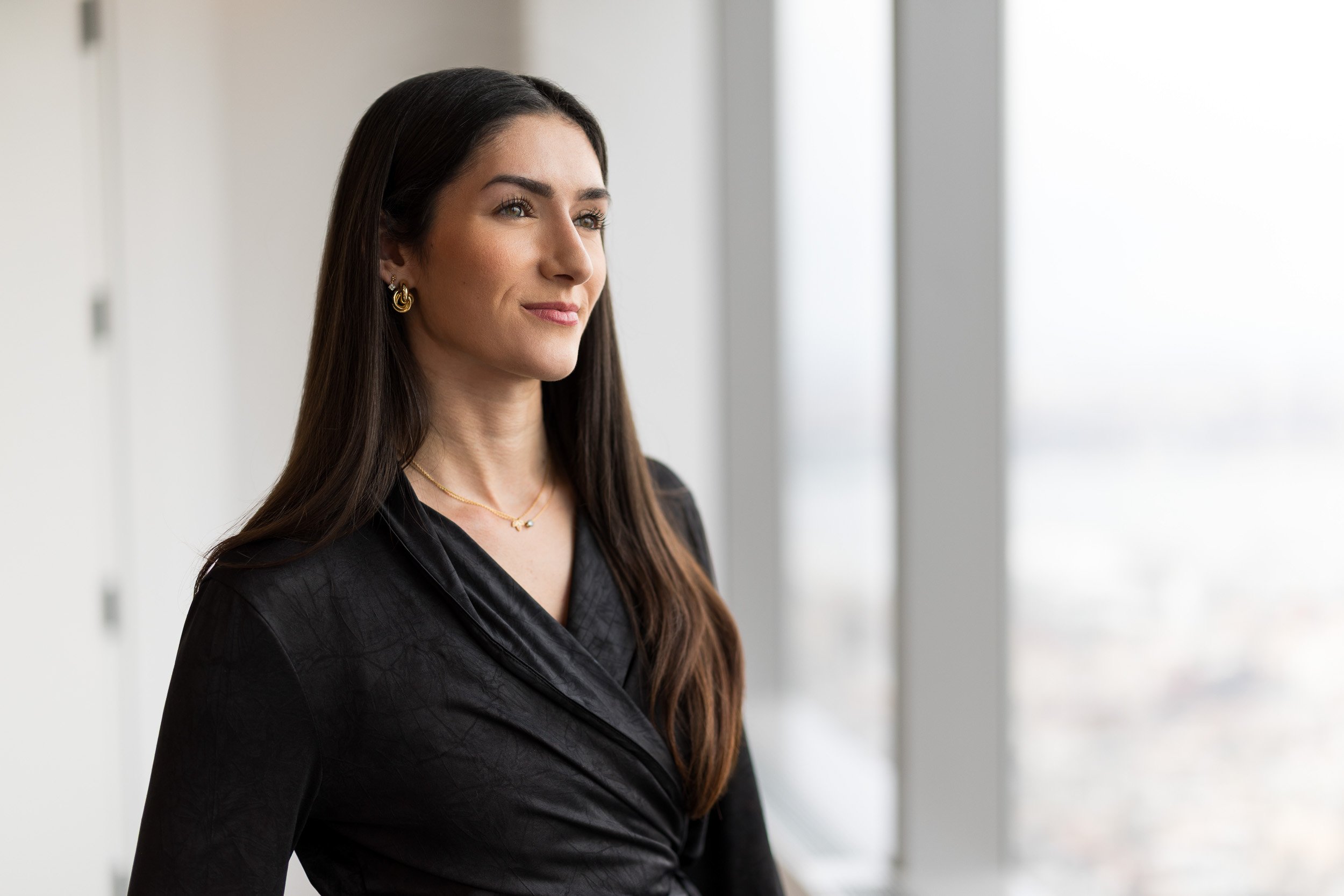 Corporate headshot of a woman in a black blouse looking off camera in an office environment with windows in the background