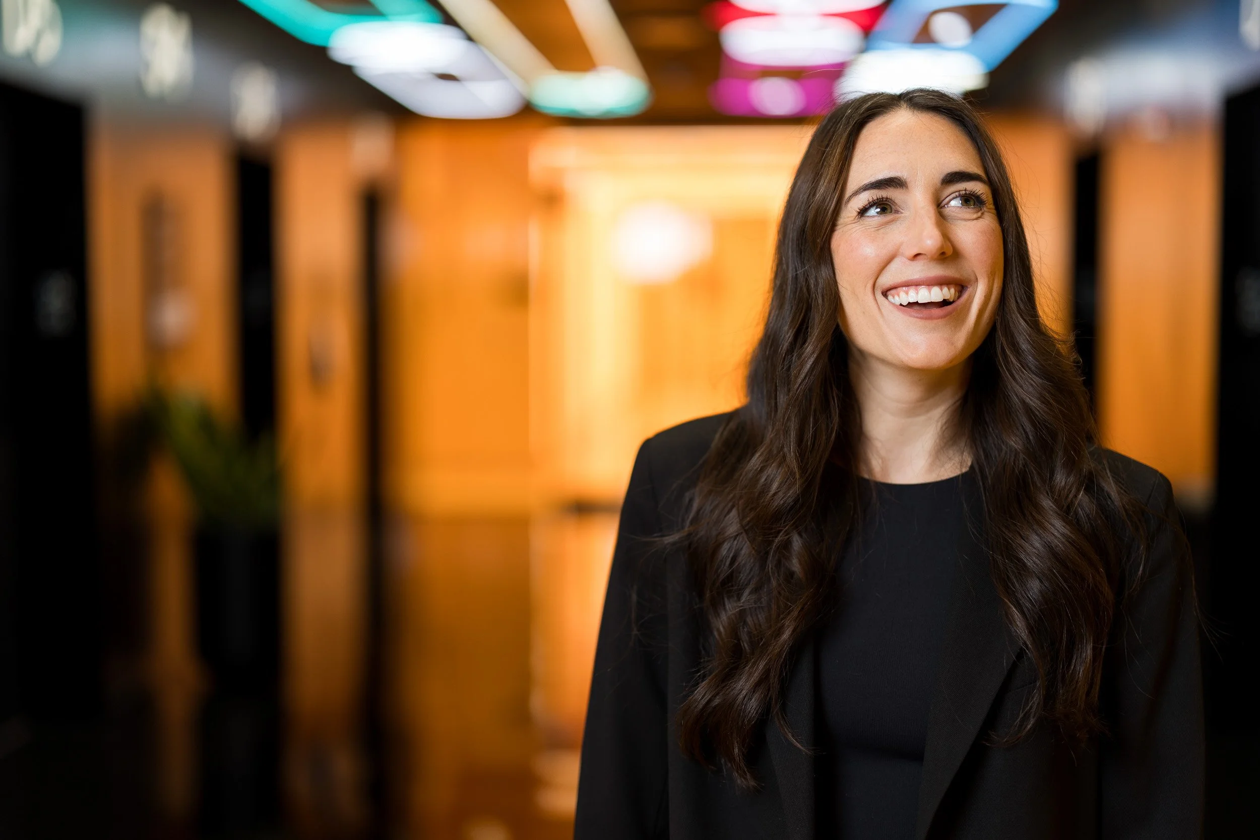 Creative corporate headshot of a smiling young white female employee with long brown hair in a black blazer and black blouse featuring a colorful orange blurred office background.