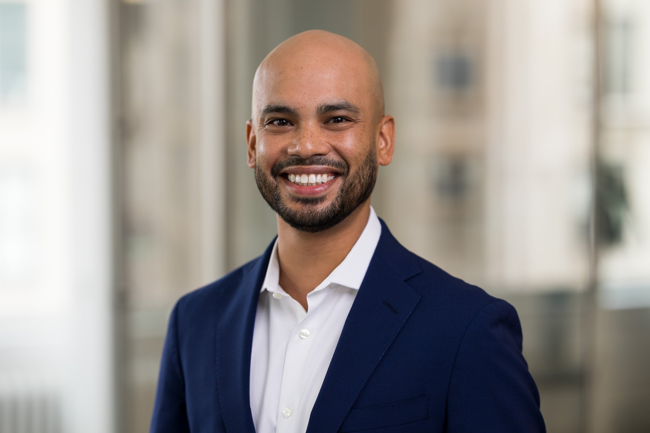 Professional headshot of a young bald male wearing a navy suit and a white shirt in his office in NYC.
