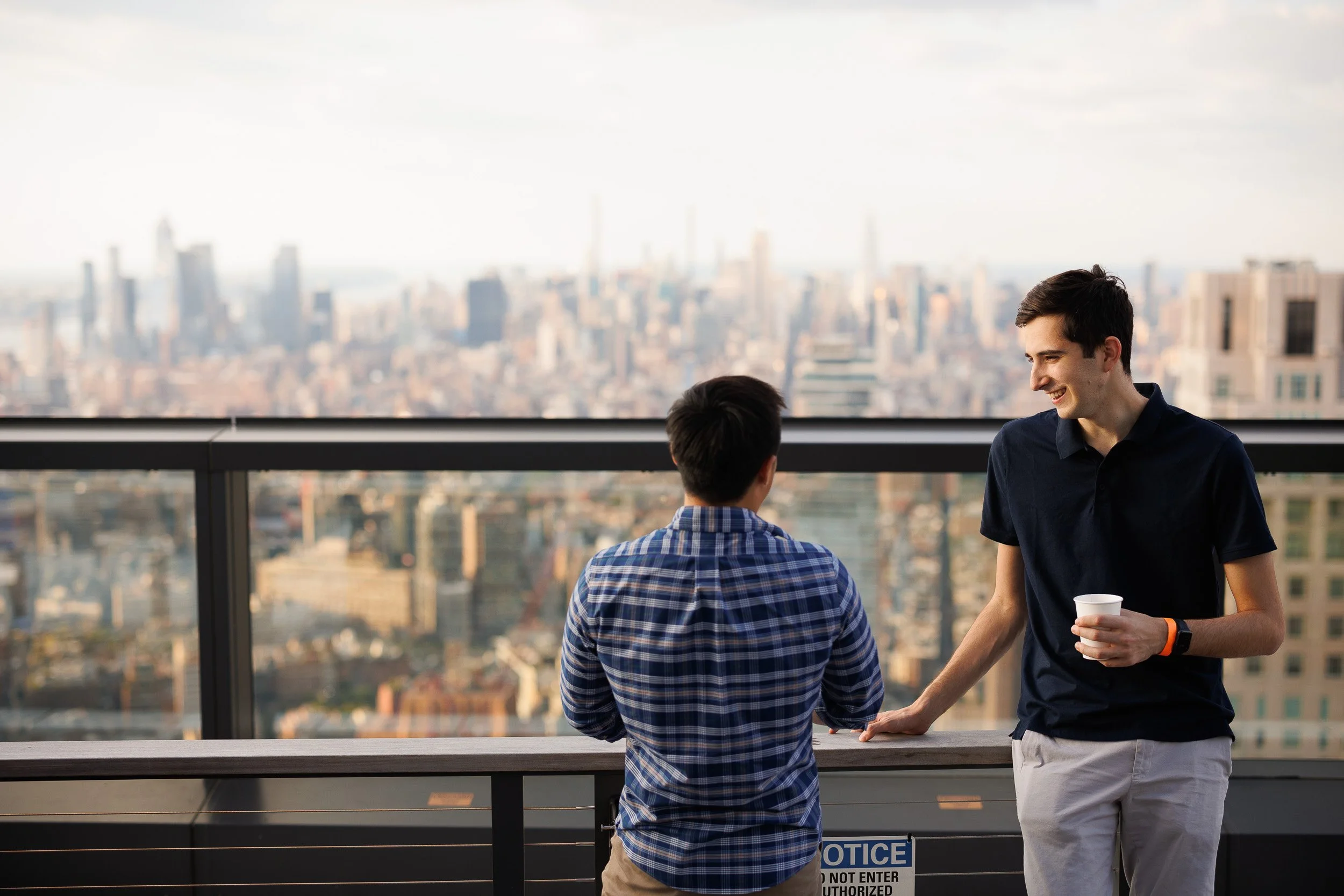 Two young men having a conversation on their office's terrace in NYC with a city skyline in the background. One is holding a cup, and they are smiling.
