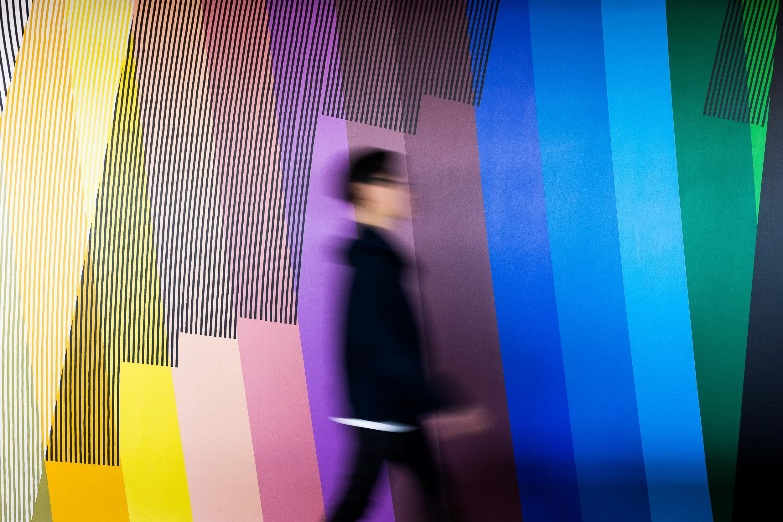 Corporate lifestyle photography of a motion-blurred male walking past a vibrant, colorful mural with vertical stripes in yellow, pink, purple, blue, and green.