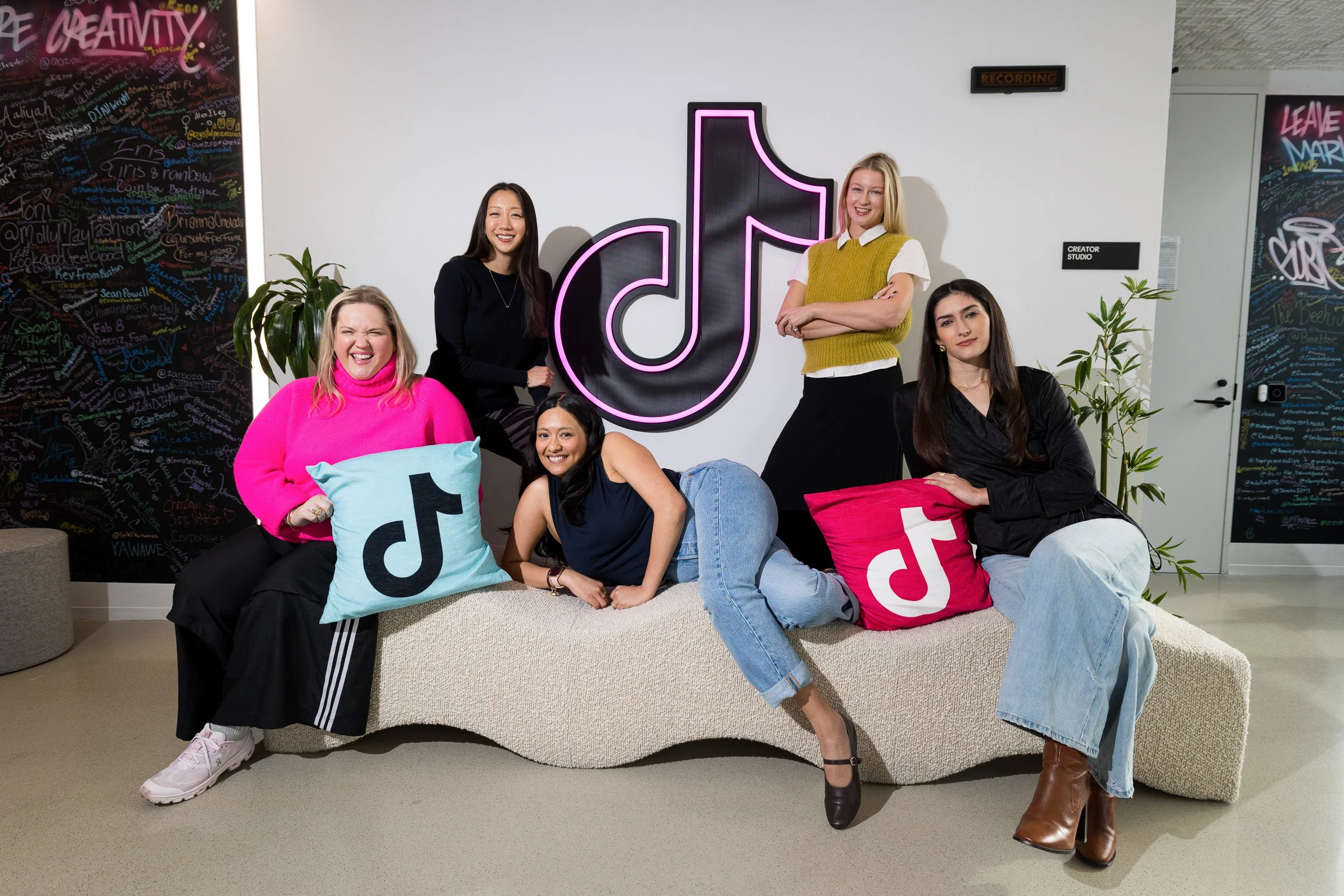 Group portrait of 5 young female employees of TikTok posed on and around a sofa with the TikTok logo centered on the wall behind them