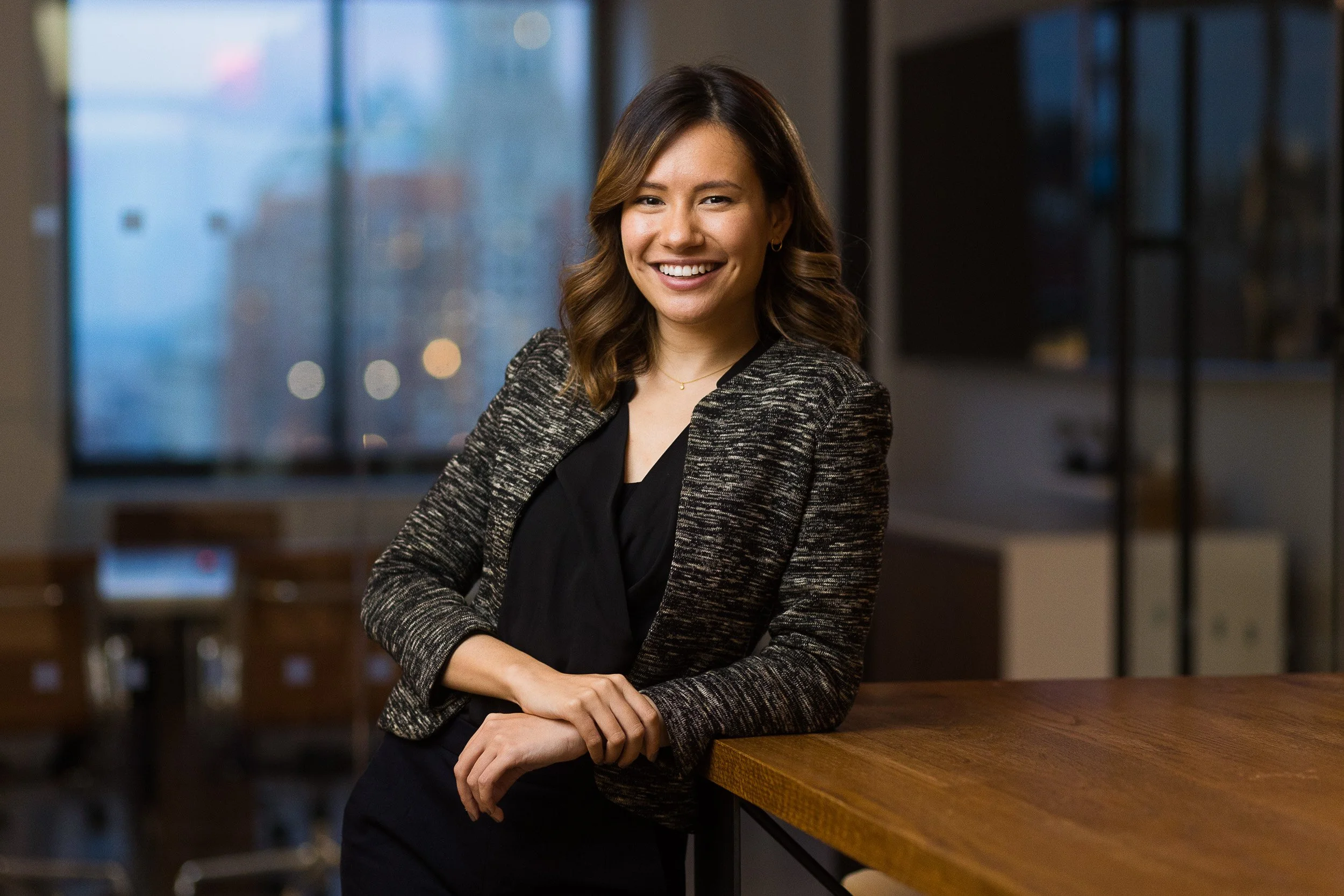 Professional headshot of a young Asian female leaning against a table in an office