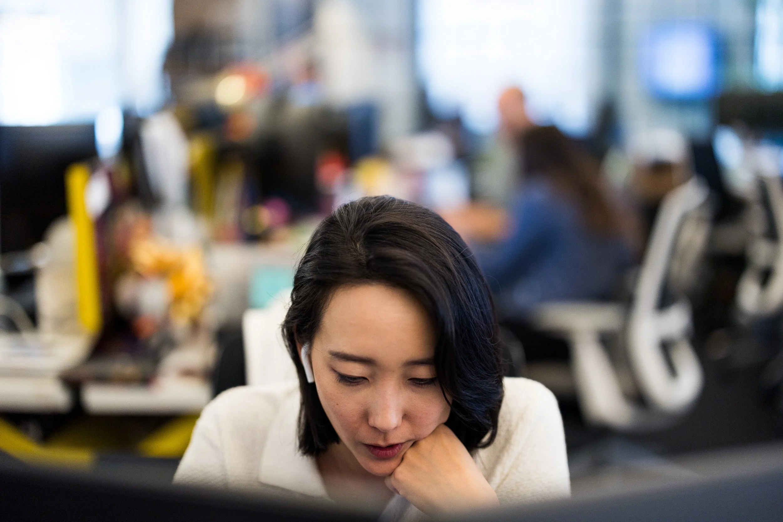 Corporate lifestyle photography of a close up shot of a young Asian female engrossed in her work in front of two computer monitors with a busy office scene in the background.