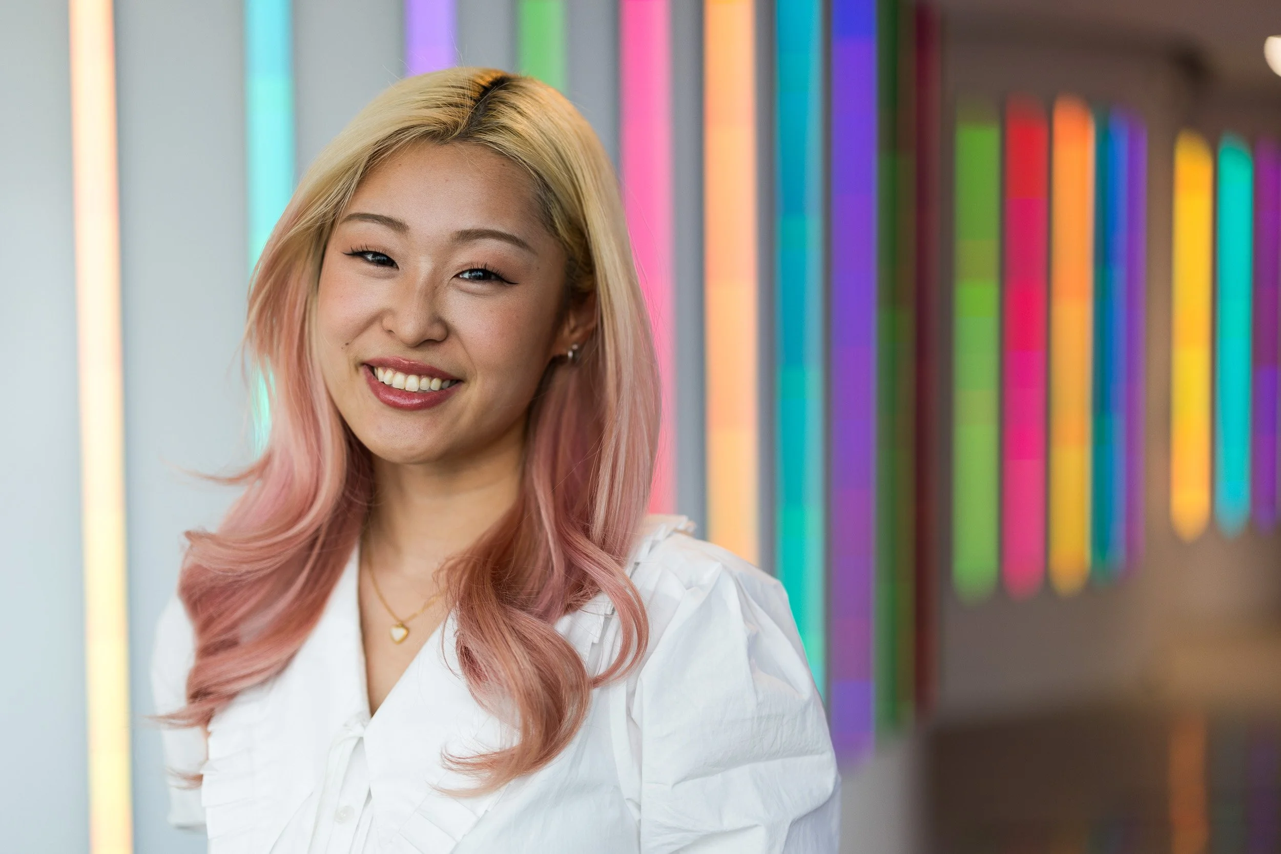 Creative corporate headshot of a young Asian female employee with blonde and pink hair smiling against a background of colorful neon lights