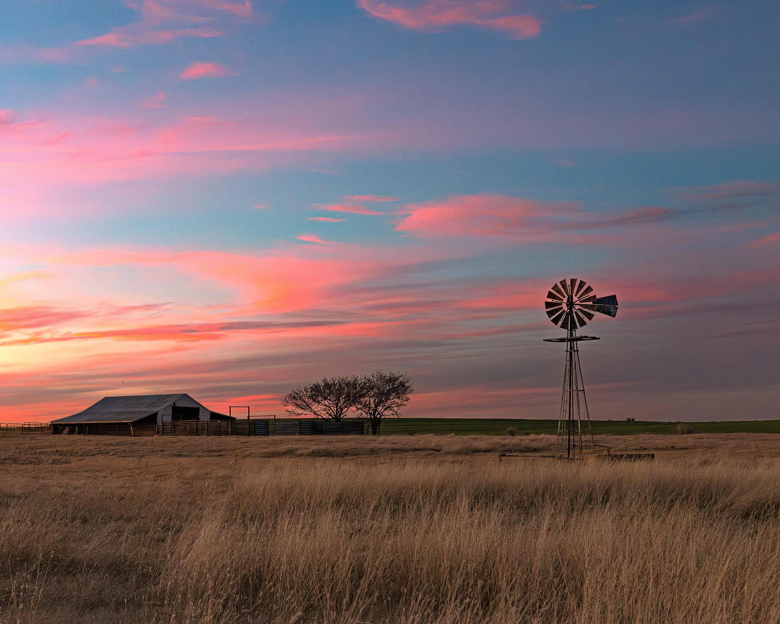 Oklahoma windmill at sunset with pastel pink clouds, prairie grass, rural farmland, and western ranch landscape on the Great Plains.