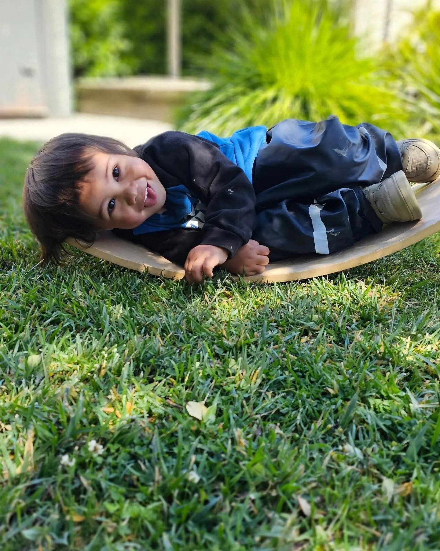 Young boy lying on his side on a wooden skateboard on green grass, smiling and looking at the camera.