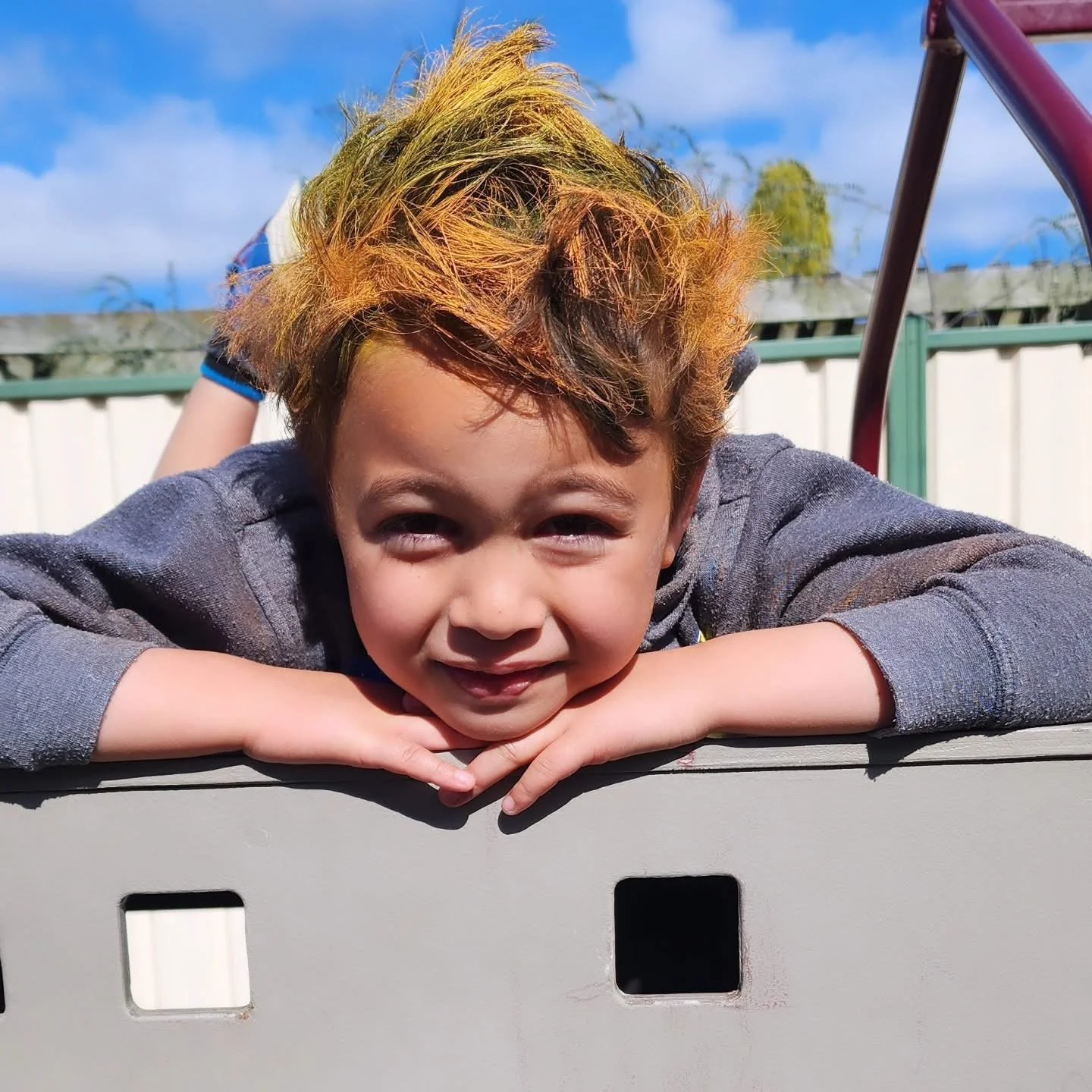 Child with tousled, colorful hair smiling and resting chin on hands, playing outdoors on a playground.
