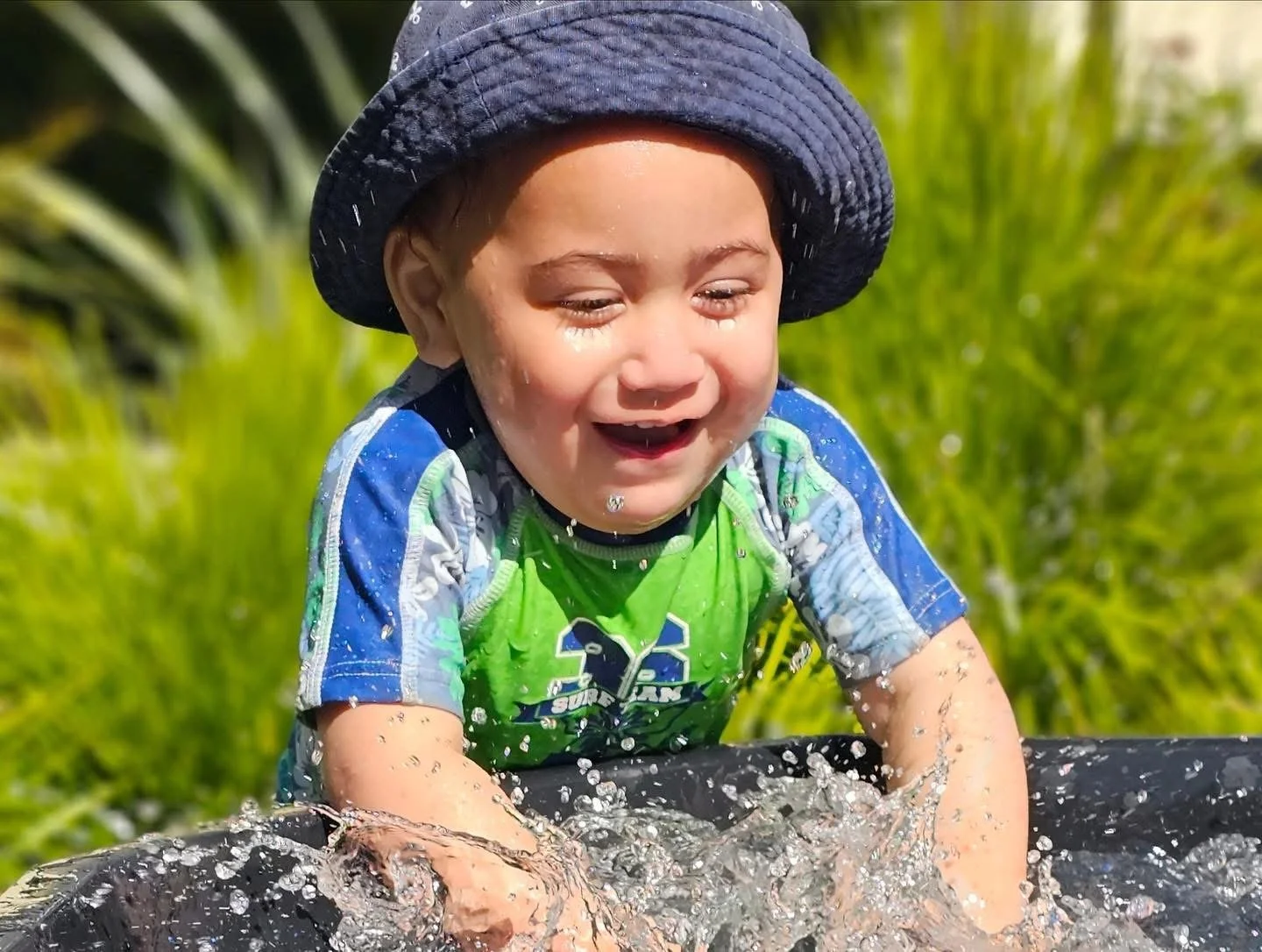 A young boy in a blue and green rash guard and a black sunhat playing in water outdoors, smiling as water splashes around him.