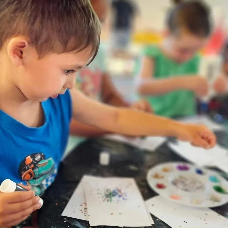 Children engaged in arts and crafts, working on colorful paintings and gluing supplies at a table.