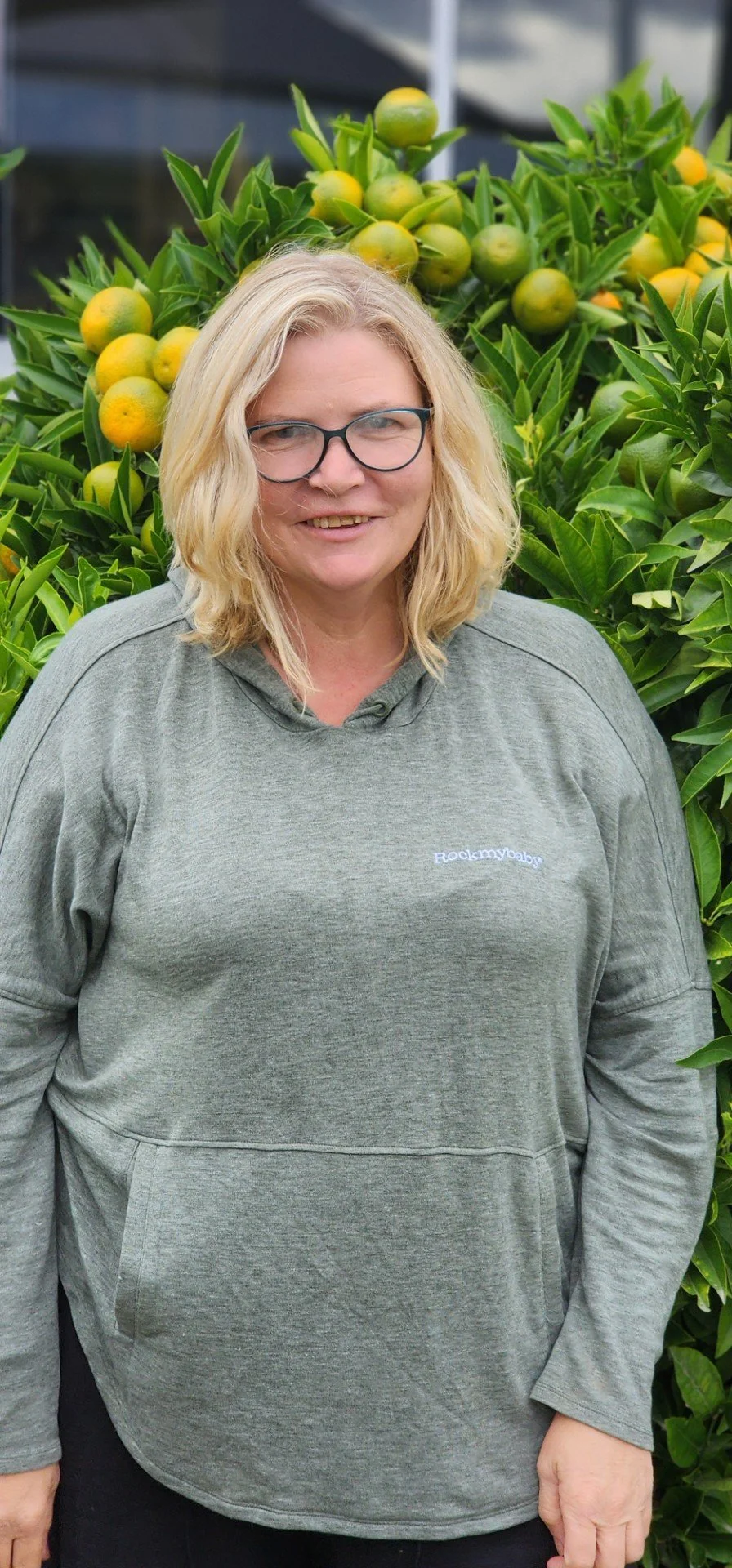 A woman with blonde hair and glasses posing outdoors in front of an orange tree with ripening oranges, wearing a gray hoodie.