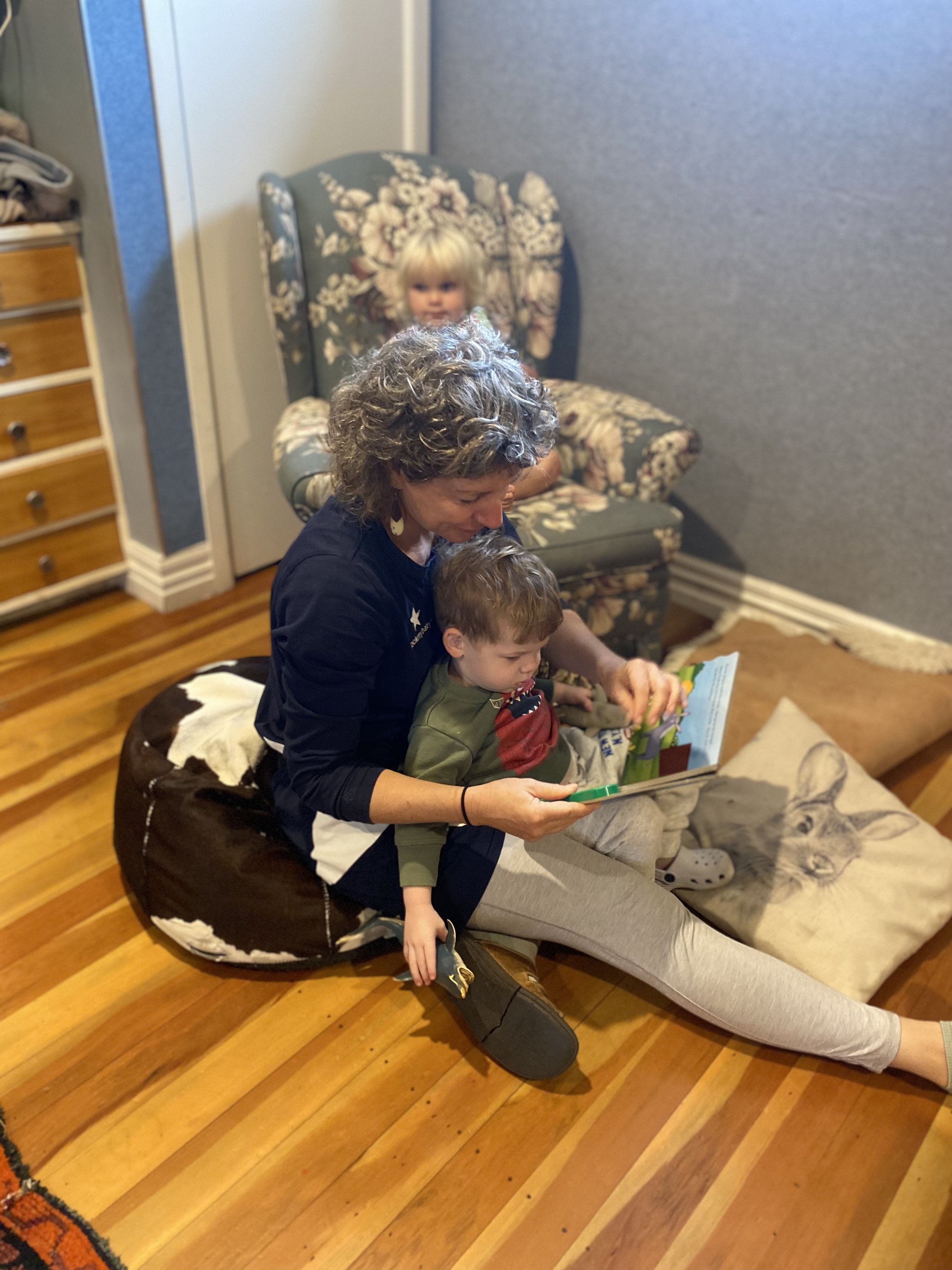 A woman with curly gray hair and two children sitting on a floor, reading a children's book. A young girl with blonde hair is sitting in a floral armchair in the background.