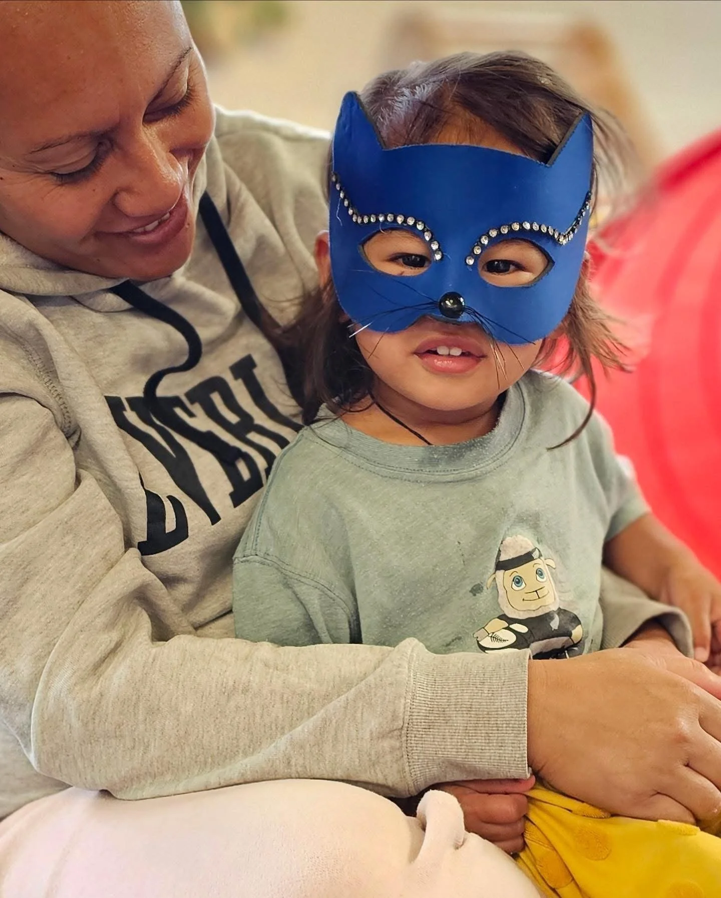 A woman holding a young girl wearing a blue cat mask decorated with rhinestones and black whiskers.