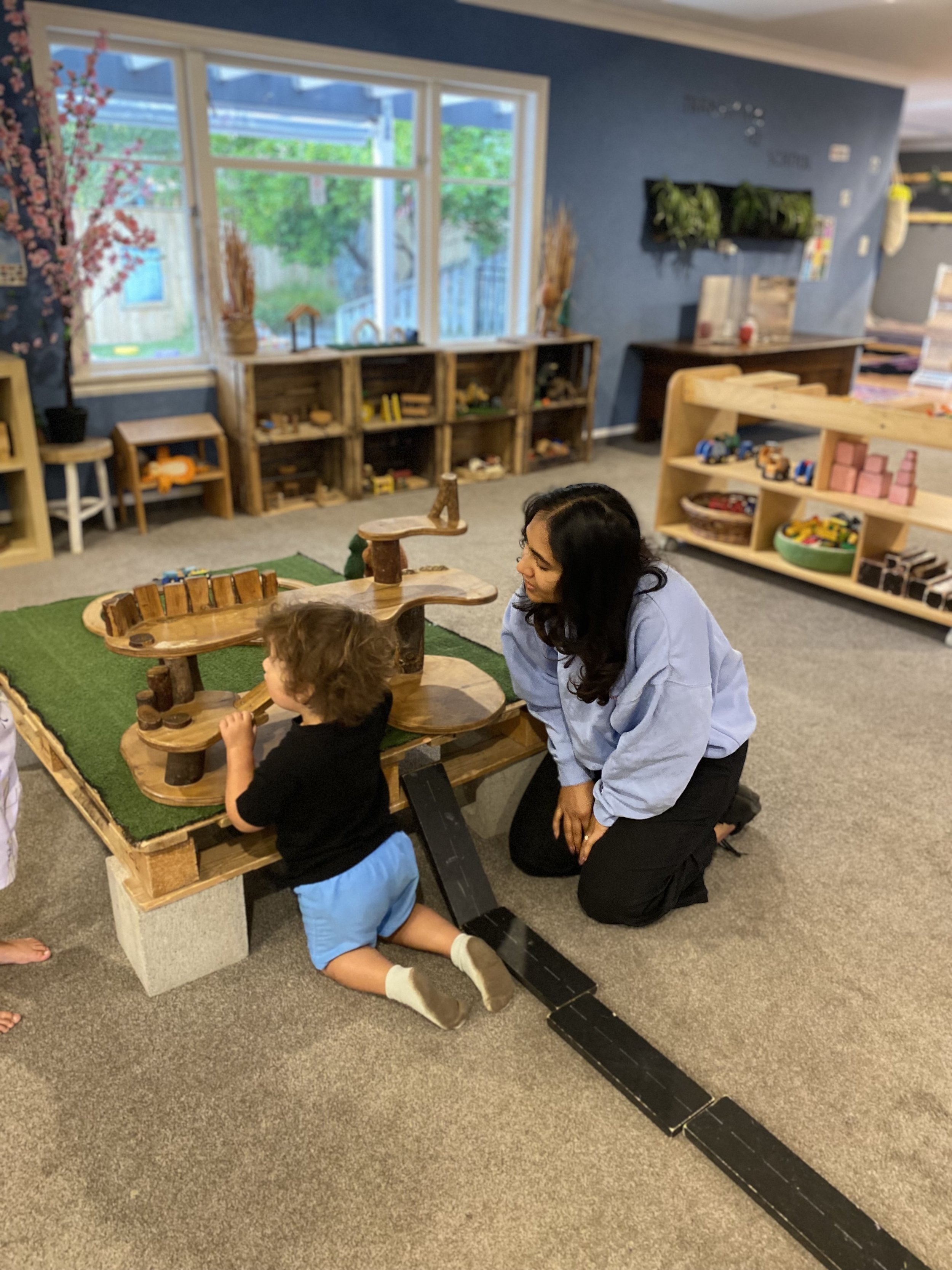 A young child and a woman playing at a wooden sandbox in a classroom with shelves and large windows.