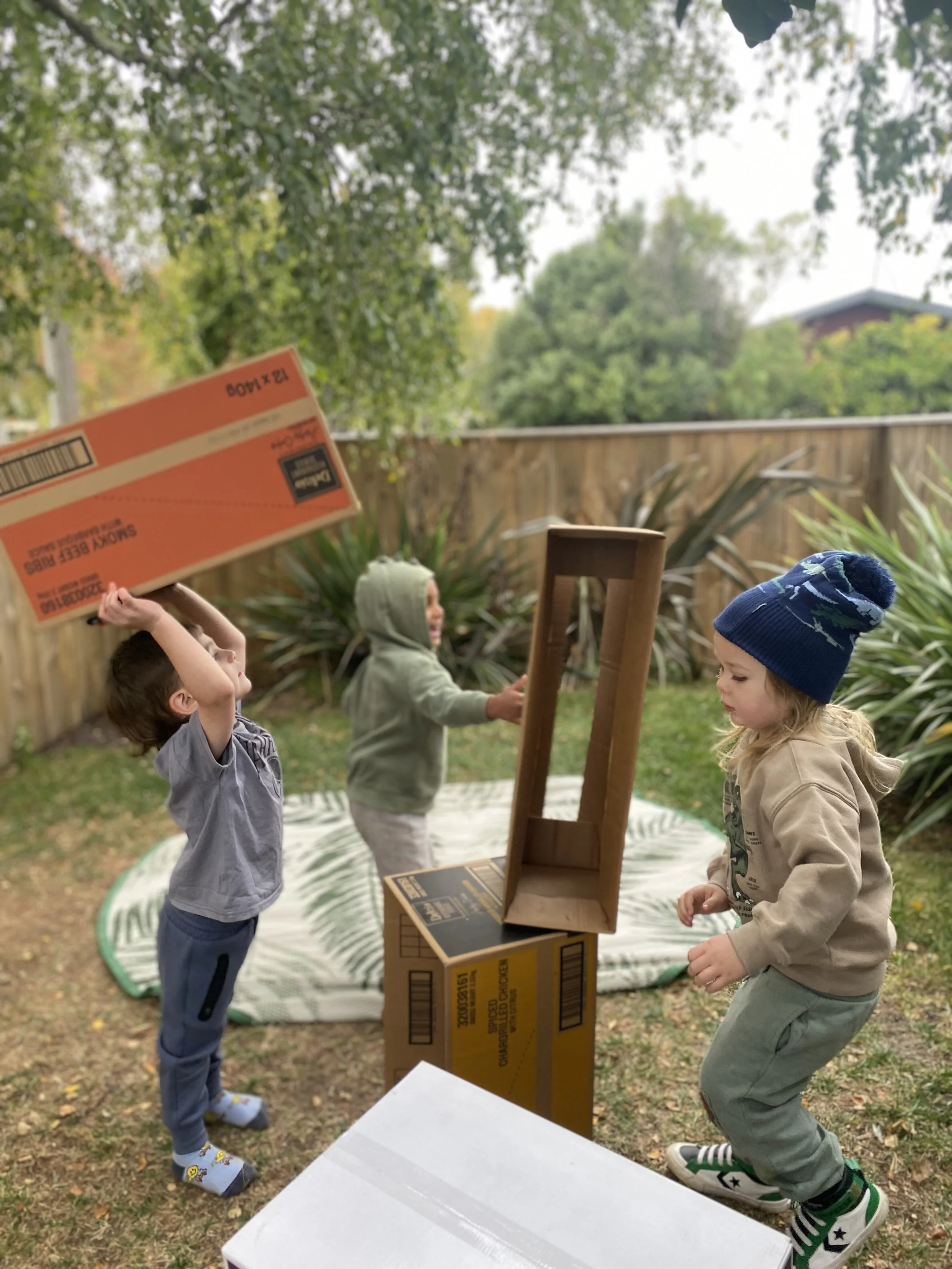 Three children playing outdoors with cardboard boxes and a wooden structure in a backyard.