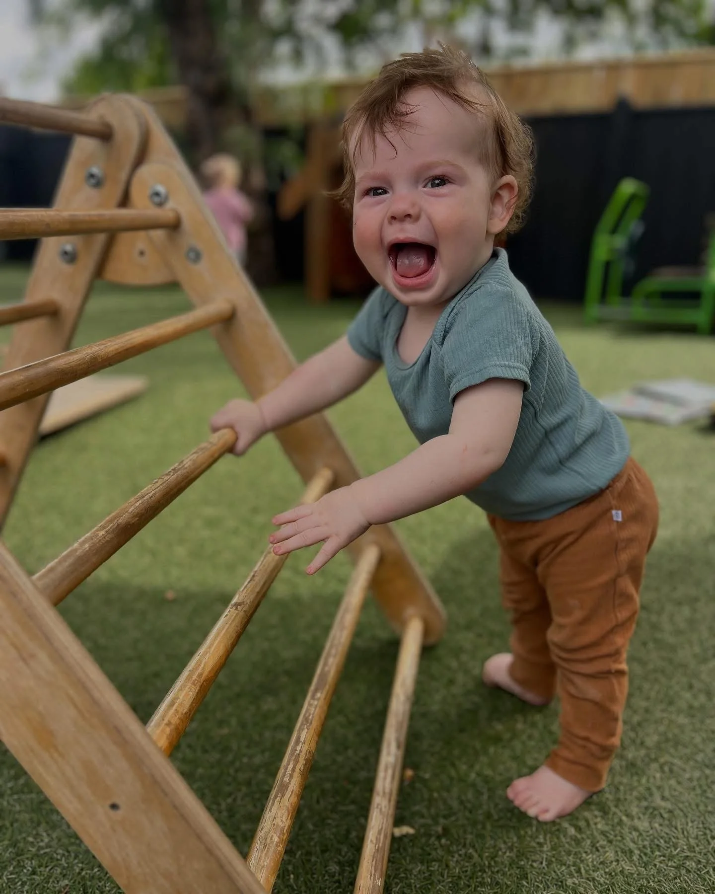 A young child with curly red hair, wearing a green t-shirt and brown pants, is outdoors on a grassy area, holding onto a wooden climbing ladder and smiling or laughing.