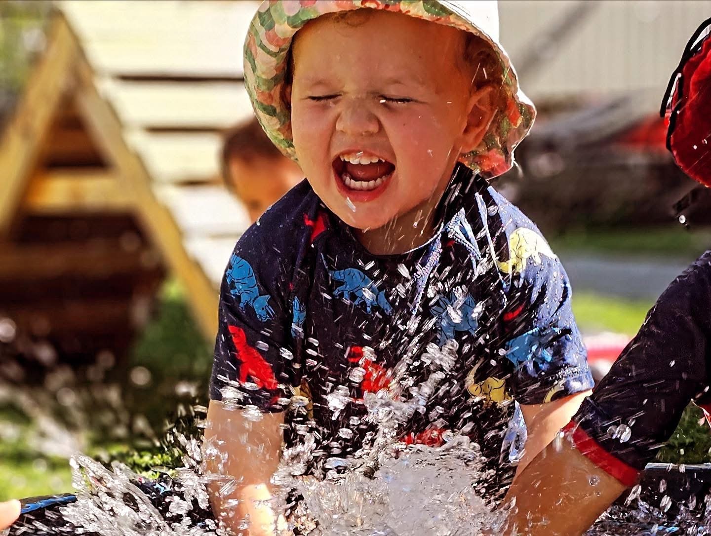 A young child with a colorful sun hat and a blue, red, yellow, and white dinosaur-patterned shirt playing in water outdoors, splashing and having fun.