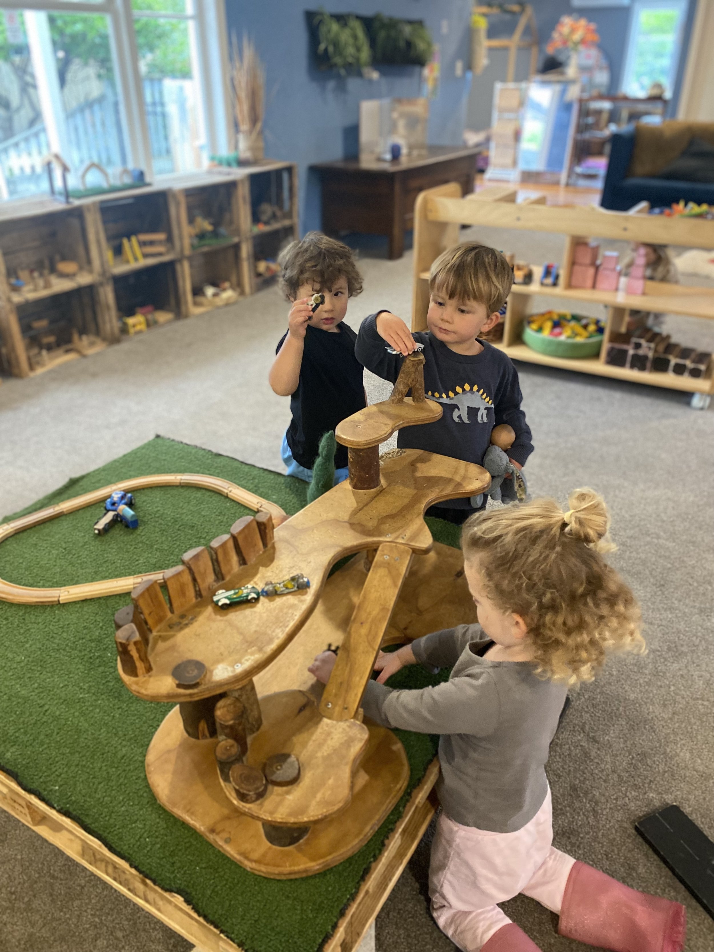 Three children playing with a wooden marble run on a green mat in a classroom or playroom.