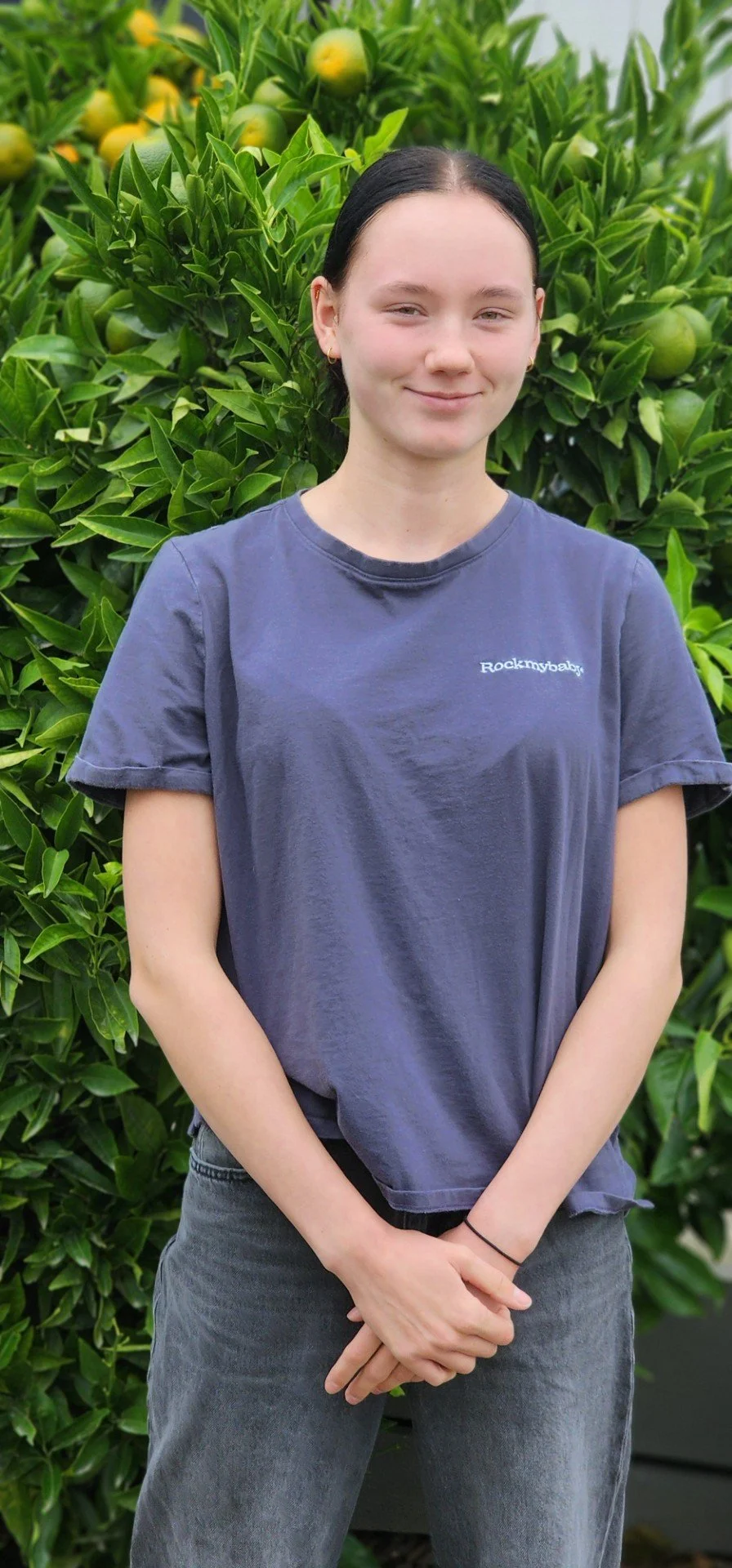 A young woman with dark hair and light skin, smiling slightly, stands in front of a lush green bush with yellow and green fruits.