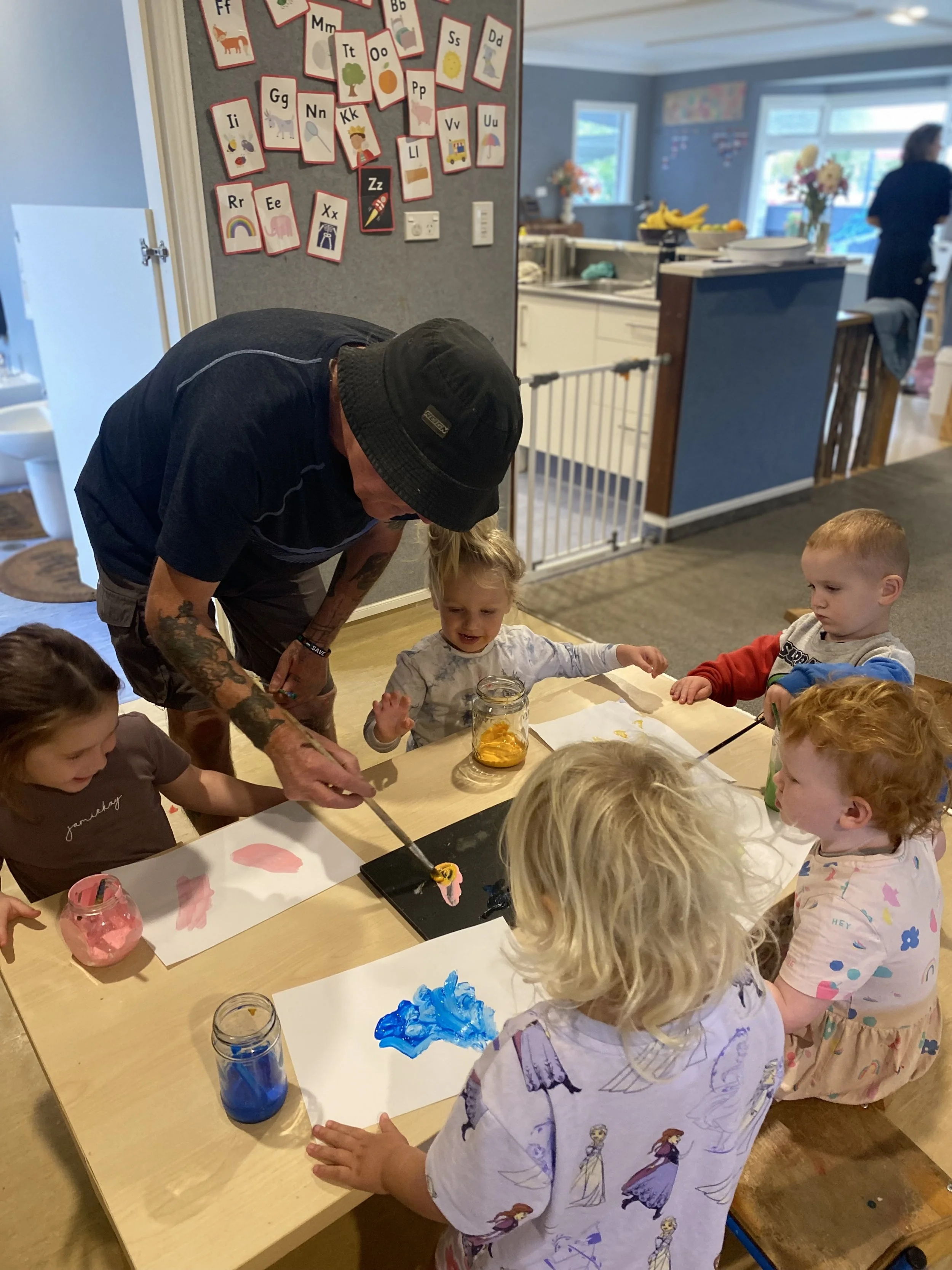 Children sitting around a table painting with help from an adult in a classroom or daycare setting.